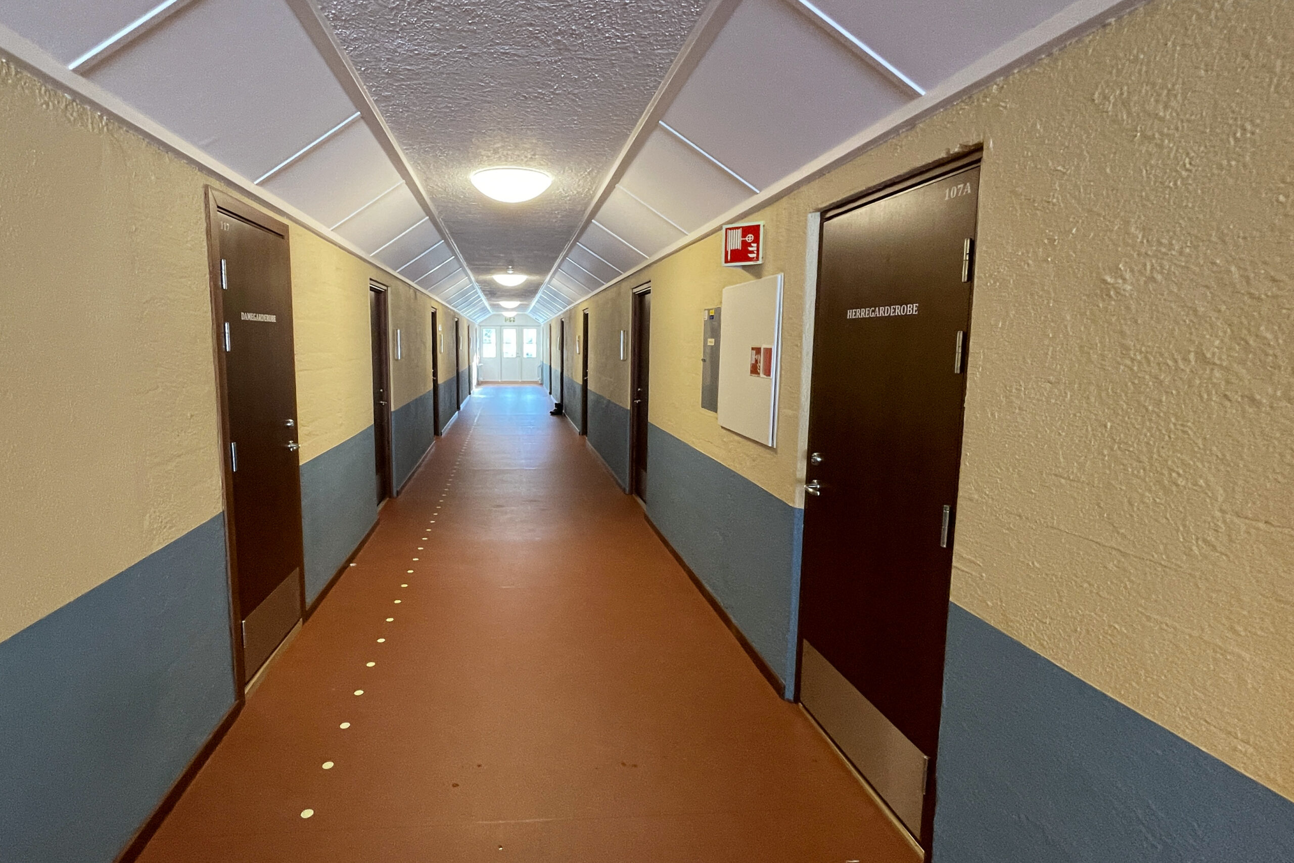 En gang med brune dører på begge sider, kremfargede og blå vegger, brunt gulv og taklampe.
