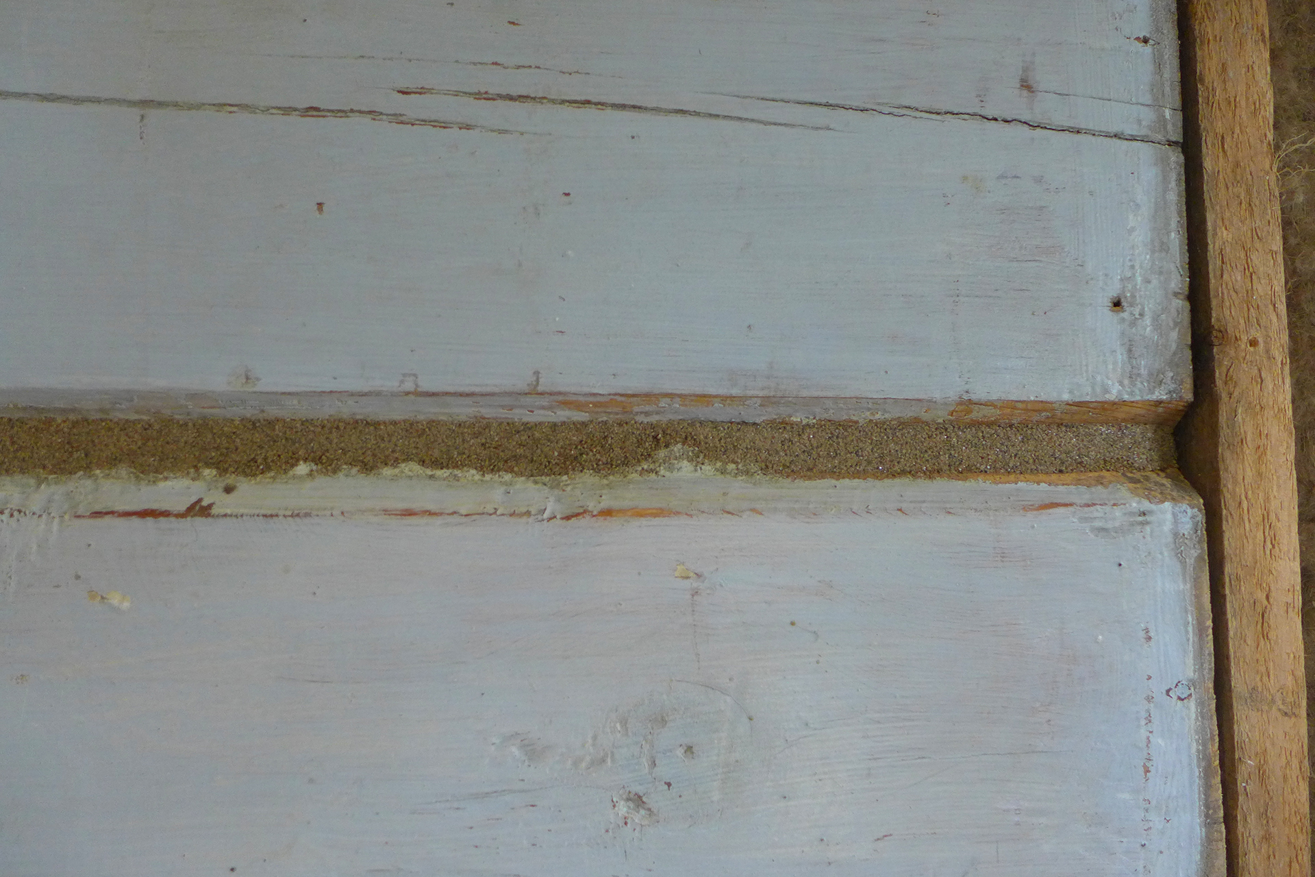 Nærbilde av en treflate med synlige sprekker, malt lyseblå, og en stripe med grov beige sparkel eller fugemasse langs en horisontal fuge.