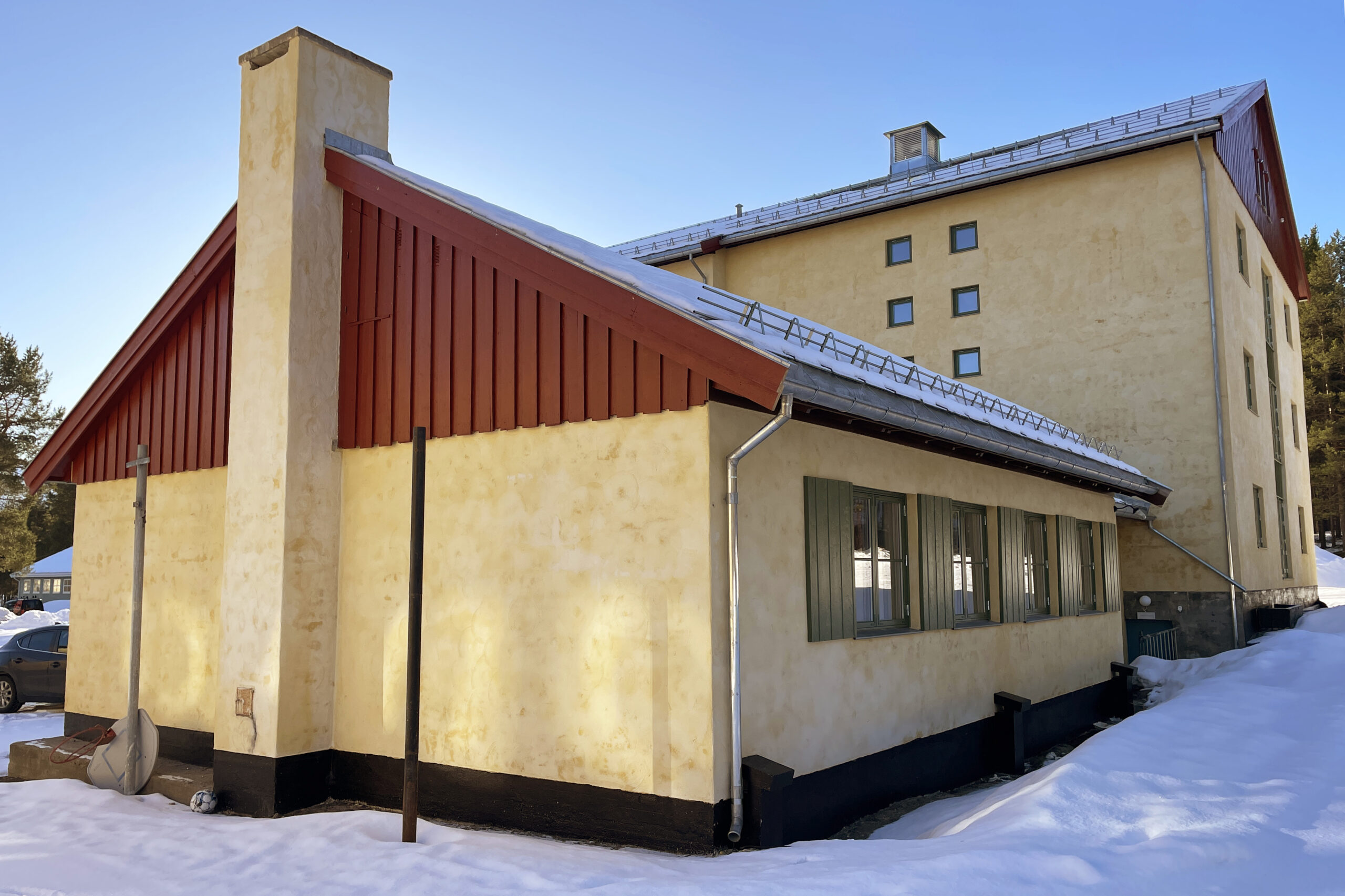 En beige bygning med rødt tak står i et snødekt område under en klarblå himmel. Bygningen har grønne vindusskodder og flere små vinduer i de øverste etasjene.