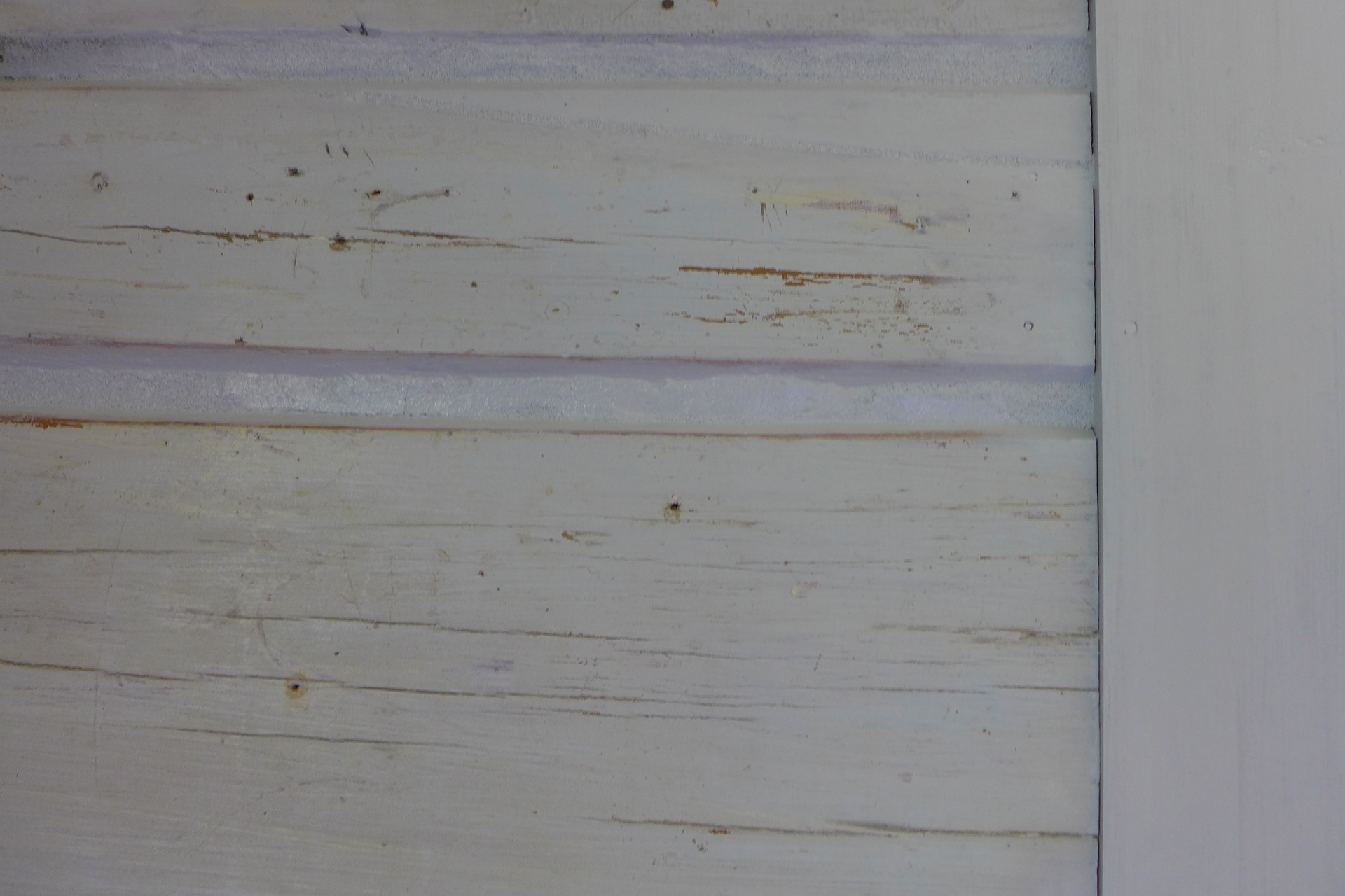 Nærbilde av en trevegg med vertikale og horisontale planker, malt lysegrå med synlig slitasje, riper og avskallet maling.