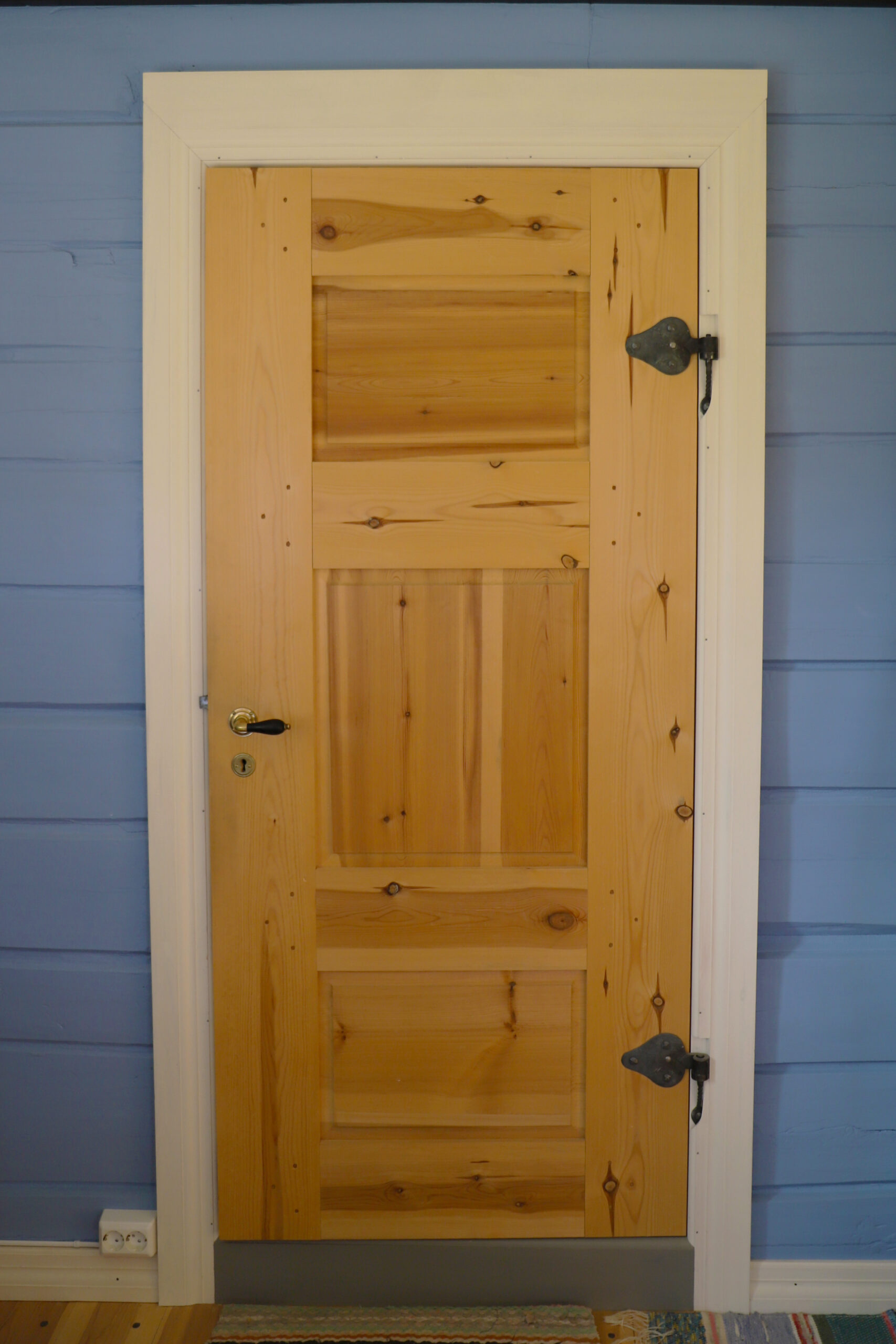 En tredør med tre horisontale paneler og hengsler i metall, satt inn i en hvit karm mot en blåmalt vegg.