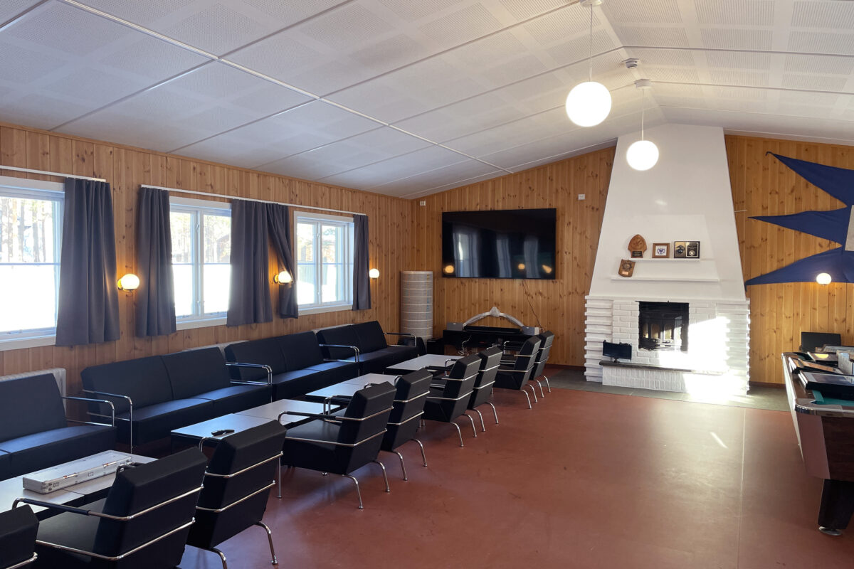 Et oppholdsrom med svarte sofaer, flere stoler, en stor veggmontert TV, en hvit mursteinspeis og vegger med trepanel og runde taklamper.