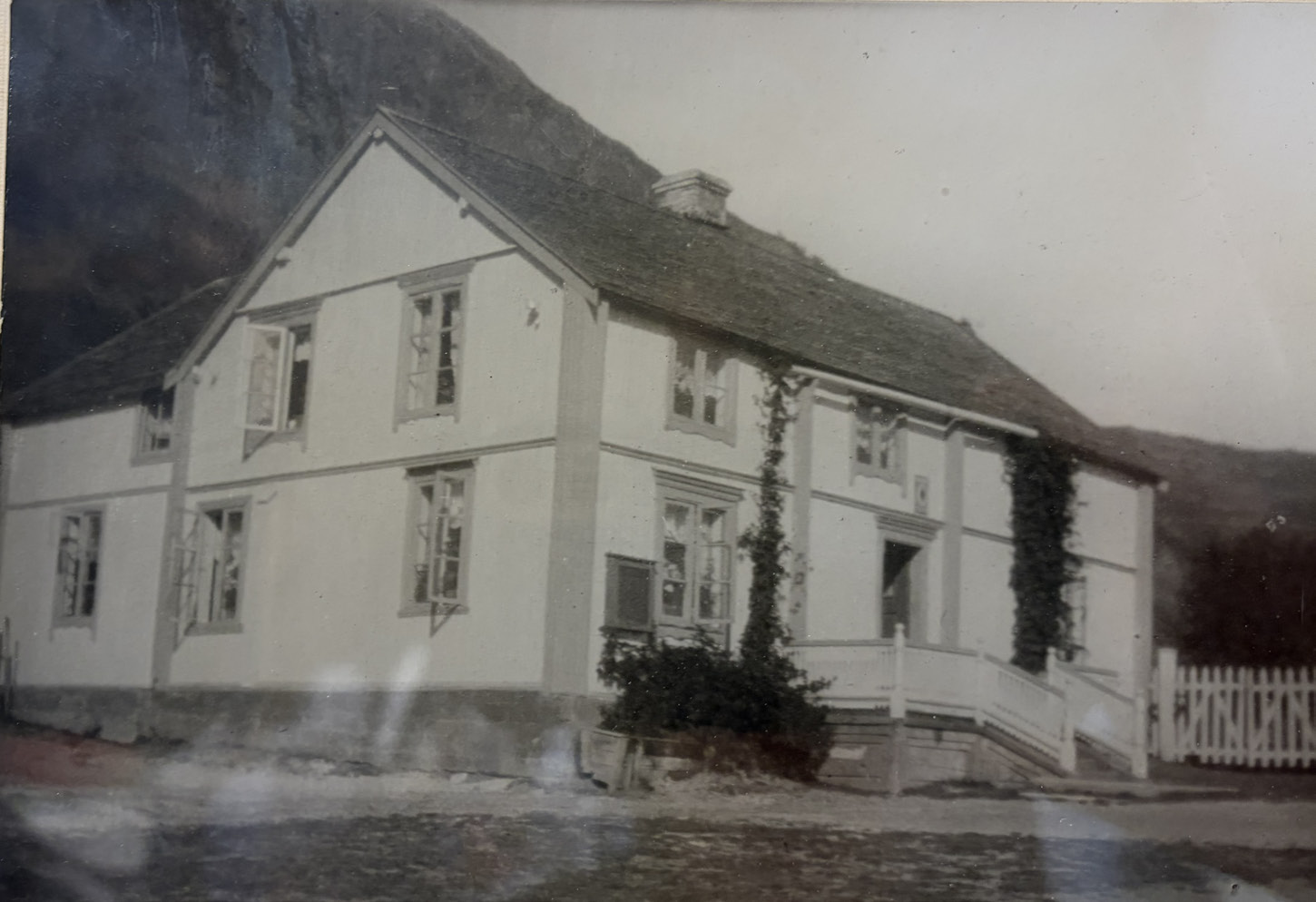 Gården Nedrum med Nergaardhuset 4 Svart-hvitt-foto av et hvitpusset hus i to etasjer med bratt tak, eføy på fasaden og et hvitt stakittgjerde, plassert mot en steinete åsside.