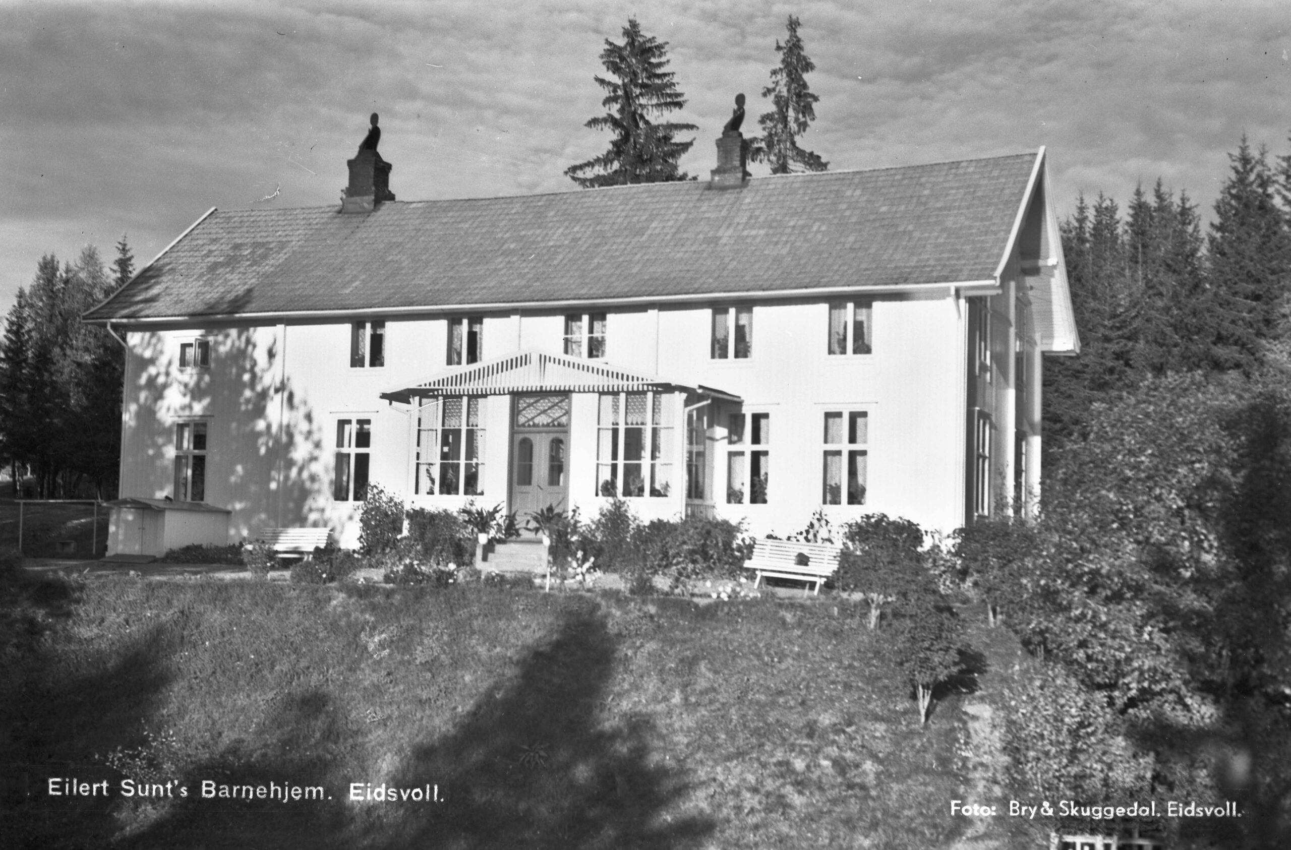 Svart-hvitt-foto av en stor, toetasjes bygning med en sentral inngang, merket "Eilert Sunts Barnehjem, Eidsvoll", omgitt av trær og busker.