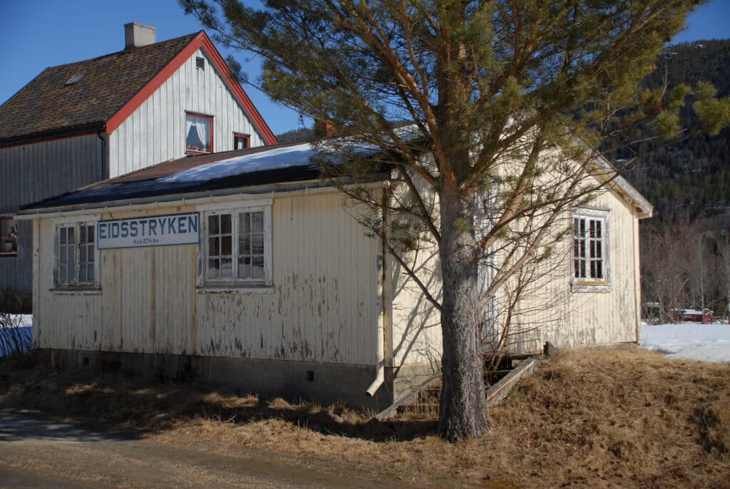 En liten, hvit, værbitt bygning med skiltet "Eidsstyrken" står ved siden av et tre, med et annet hus og snø på bakken i bakgrunnen.