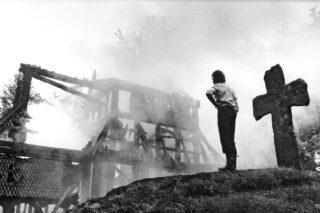 En person står på en høyde i nærheten av en stor gravstein og ser på de røykfylte ruinene av en brent trebygning med trær i bakgrunnen.