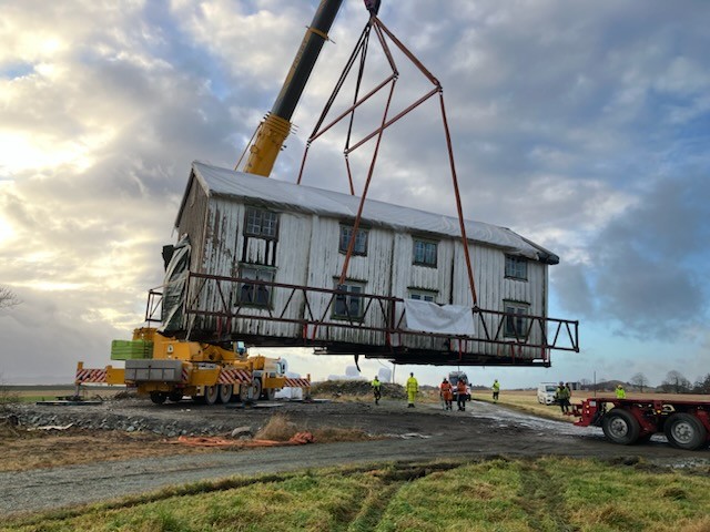 En kranbil løfter et av husene på Ørlandet, mens arbeidere i sikkerhetsutstyr observerer arbeidet fra bakken under en delvis overskyet himmel.