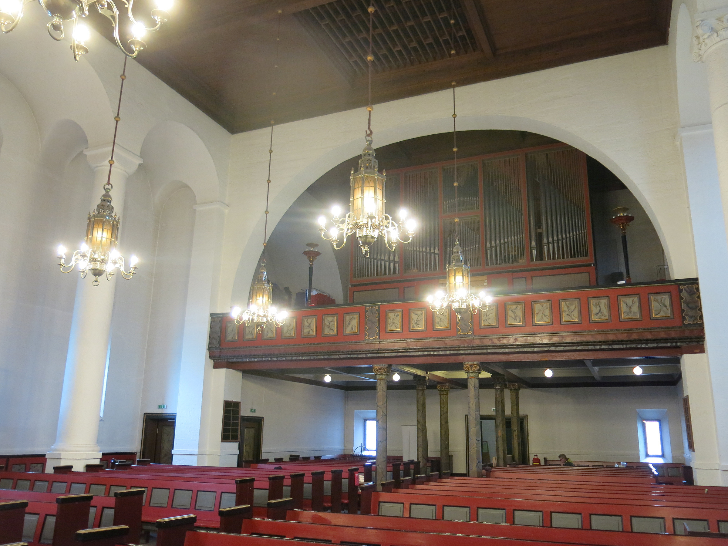 Interiør av en kirke med røde kirkebenker, utsmykkede lysekroner og et stort pipeorgel på en øvre balkong som støttes av søyler.