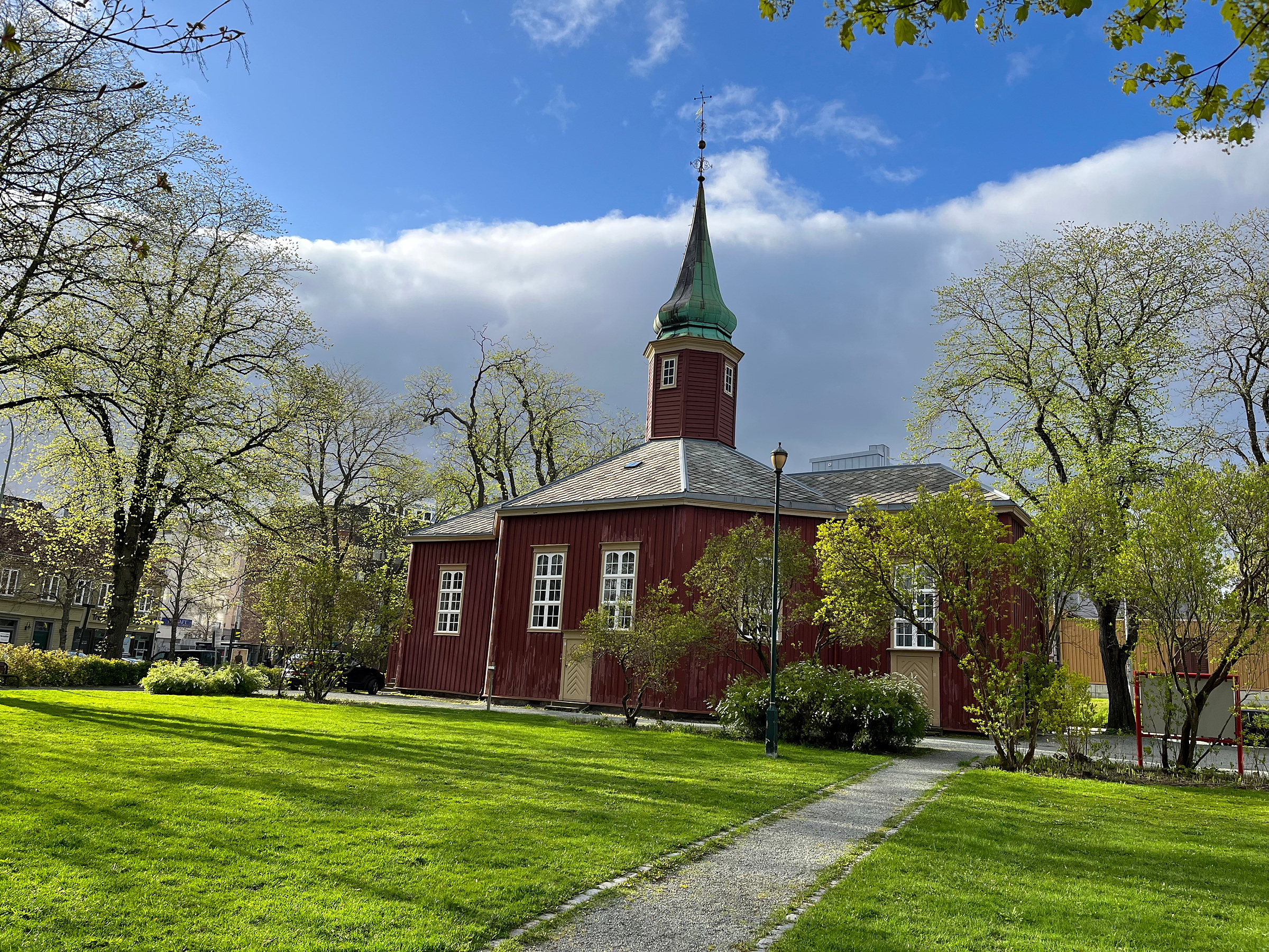 En liten rød trekirke med et grønt kirketårn står i en gressbevokst park omgitt av trær under en delvis overskyet himmel.