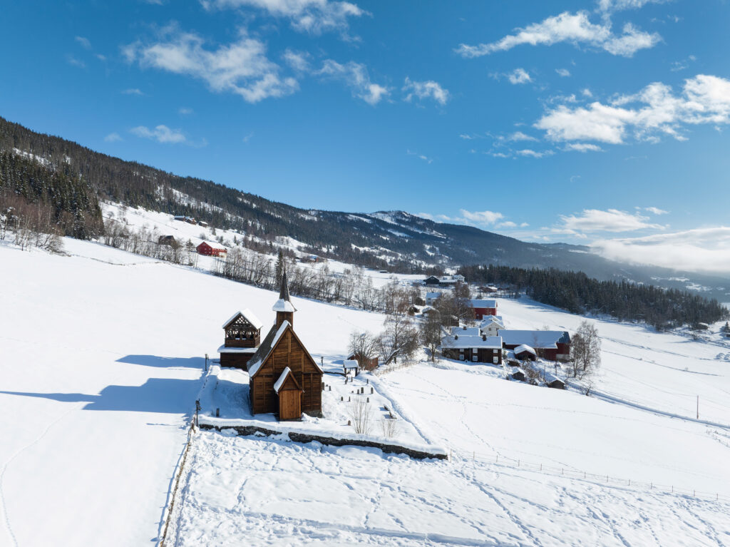 En trekirke og nærliggende bygninger ligger i et snødekt landskap med åser og trær under en delvis overskyet blå himmel.