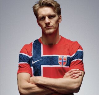 En mann med blondt hår står med armene i kors, iført en rød norsk fotballtrøye med et stort blåhvitt skandinavisk kors og det norske riksvåpenet.