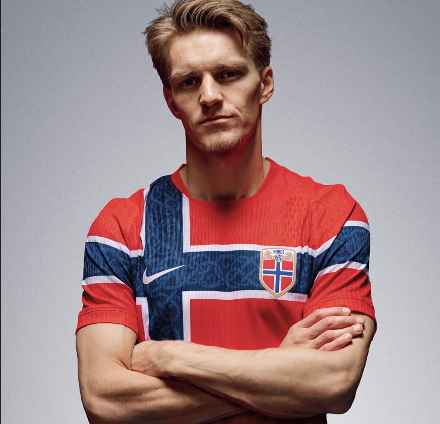 En mann med blondt hår står med armene i kors, iført en rød norsk fotballtrøye med et stort blåhvitt skandinavisk kors og det norske riksvåpenet.