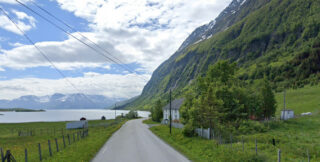 En landevei går langs en fjord med fjell i bakgrunnen; et hvitt hus og grønne trær ligger på høyre side av veien under en delvis overskyet himmel.