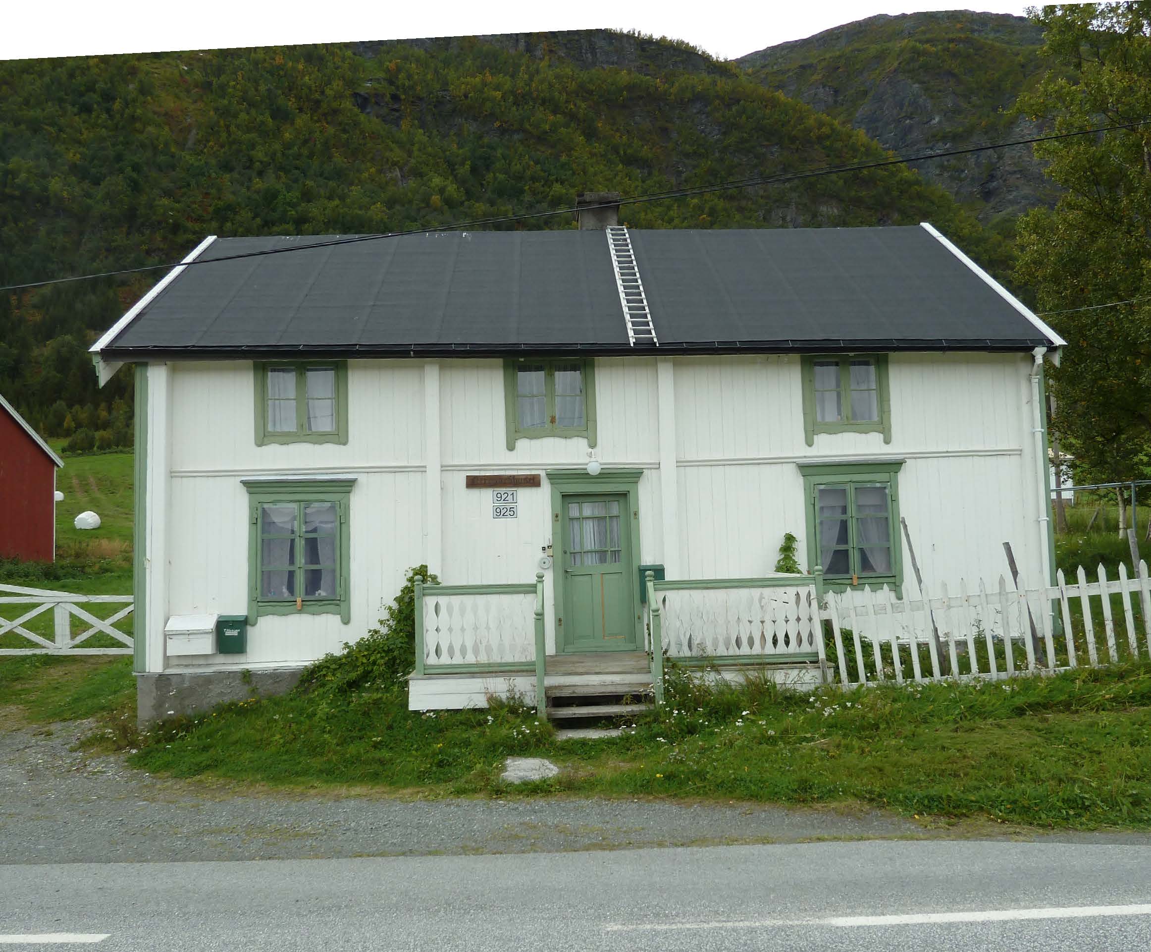 Et hvitt trehus med grønne kanter og en overbygd veranda, plassert ved siden av et stakittgjerde og et gressområde, med et fjell i bakgrunnen.