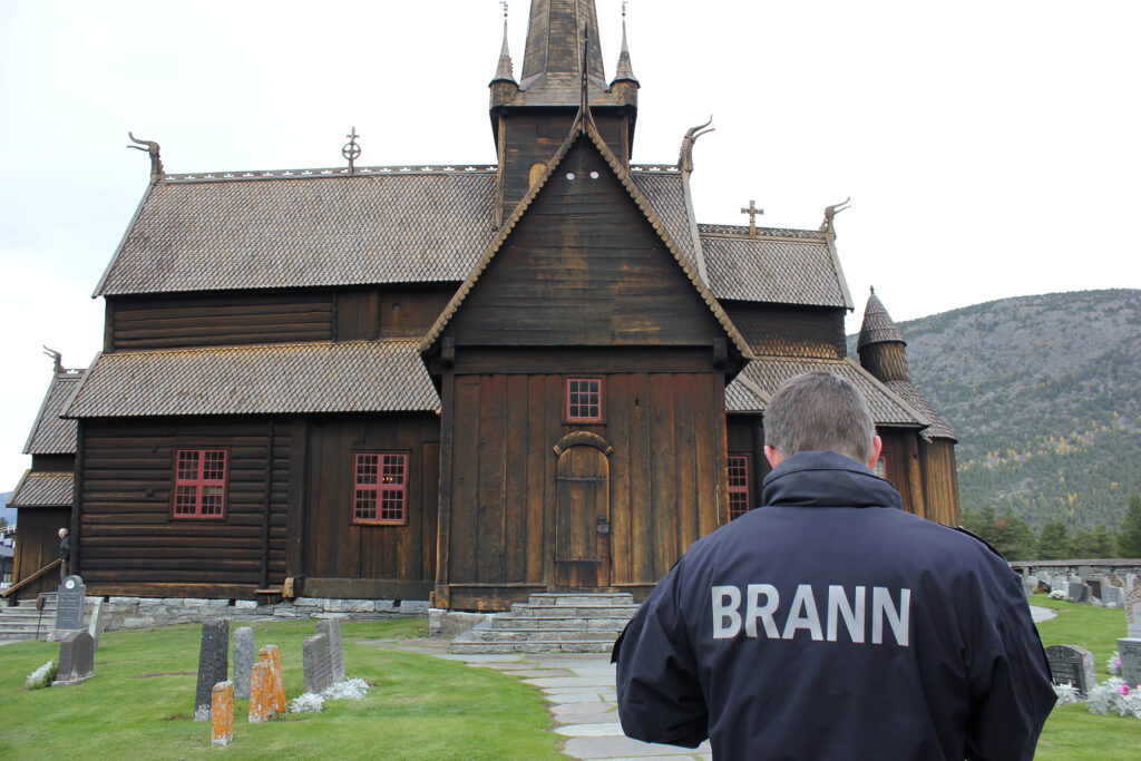 En person iført en jakke merket BRANN står foran en stor trekirke med røddekorerte vinduer og bratte, utsmykkede tak.