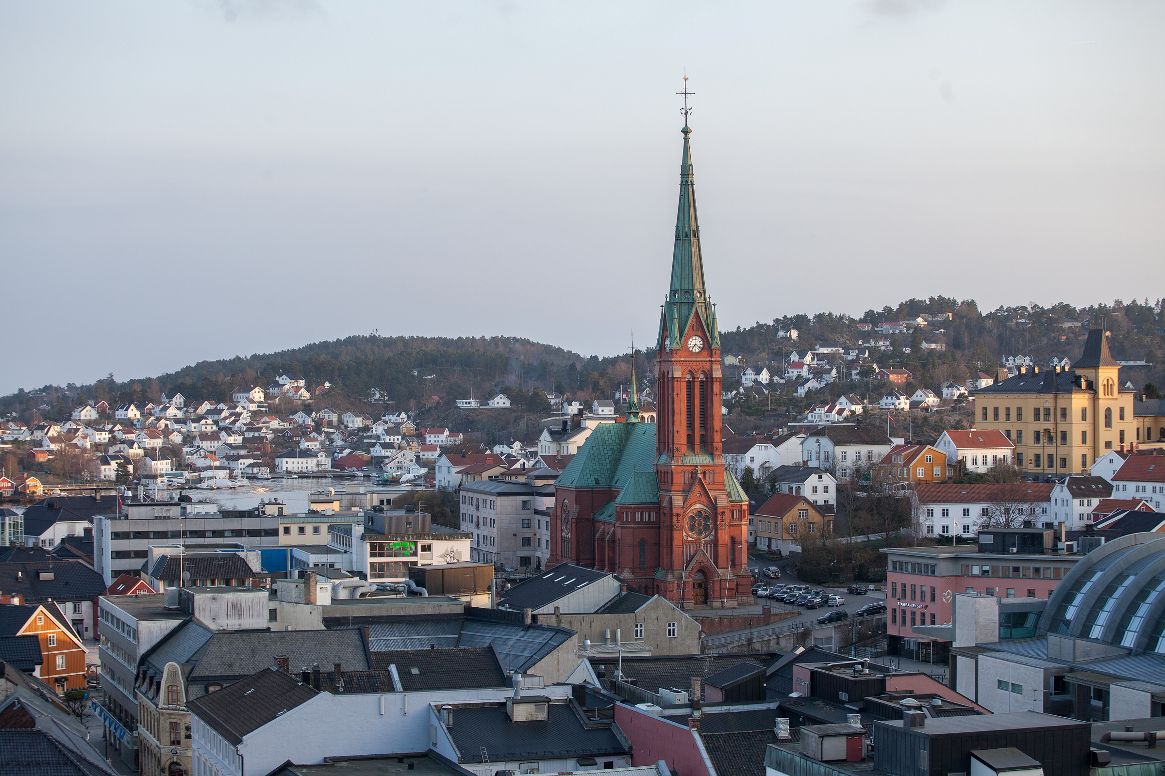 Et bybilde med en fremtredende kirke i rød teglstein med et høyt, grønt spir, omgitt av bygninger og hus på bølgende åser under en klar himmel.