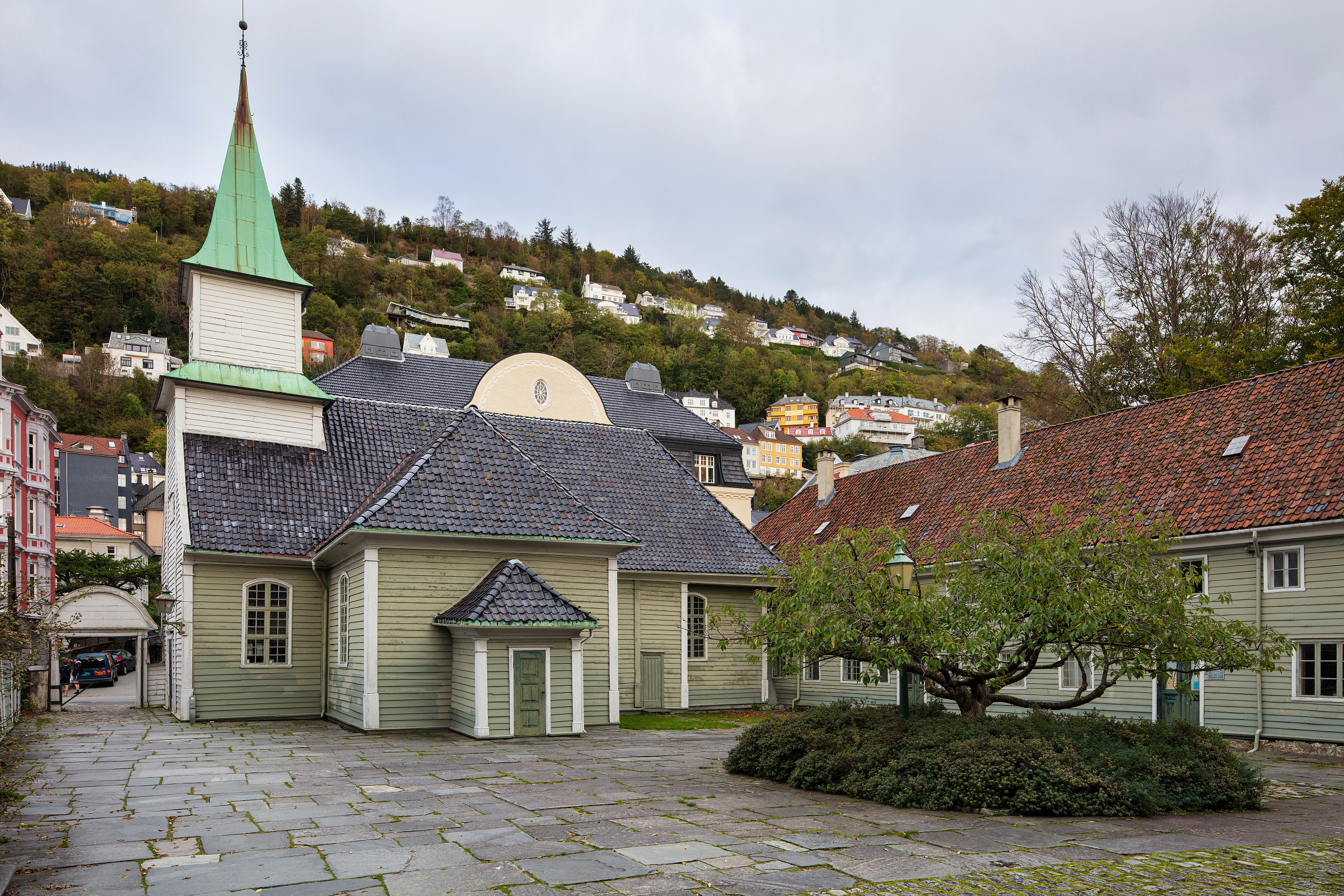 En liten trekirke med et grønt kirketårn står ved en gårdsplass av stein, med hus på en høyde i bakgrunnen under en overskyet himmel.