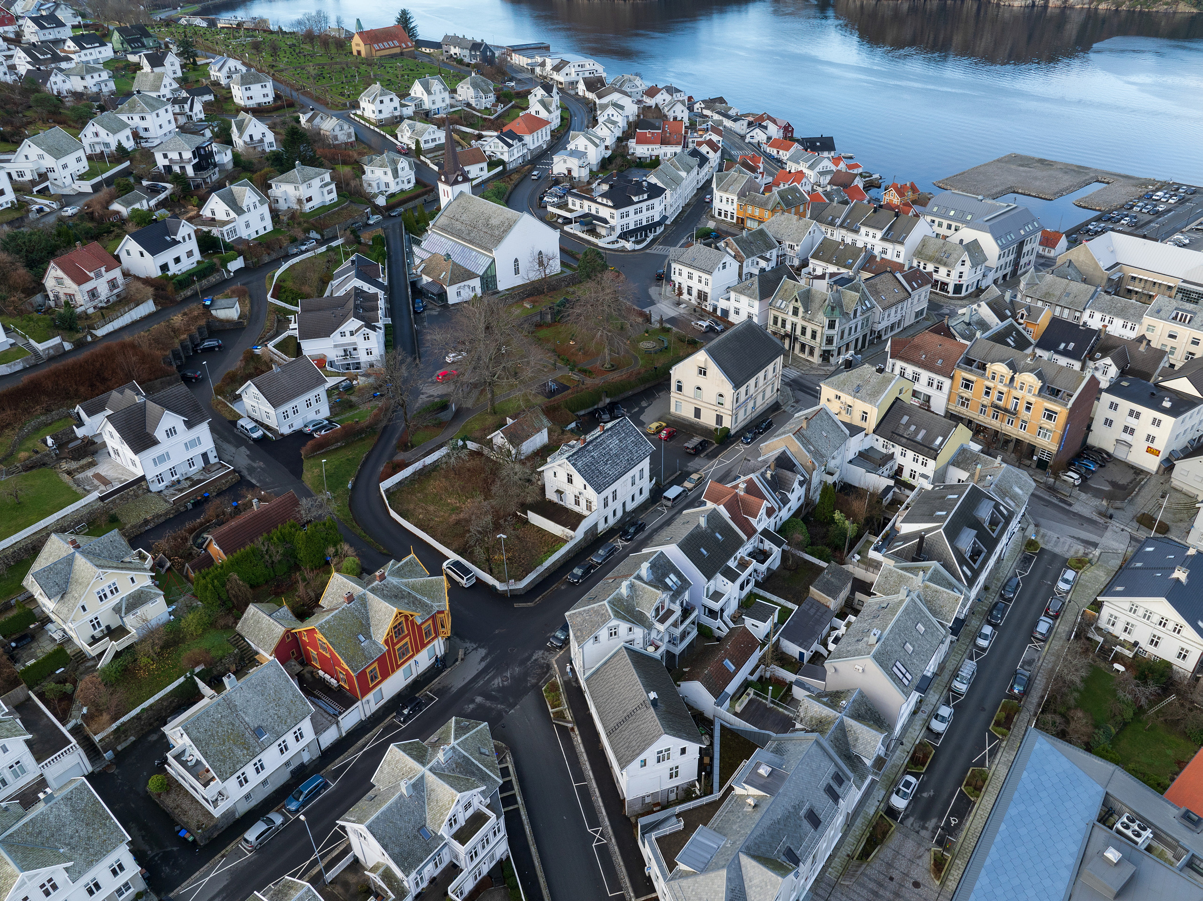 Flyfoto av en kystby med hvite hus, noen med grå tak, plassert langs svingete gater nær vannet.
