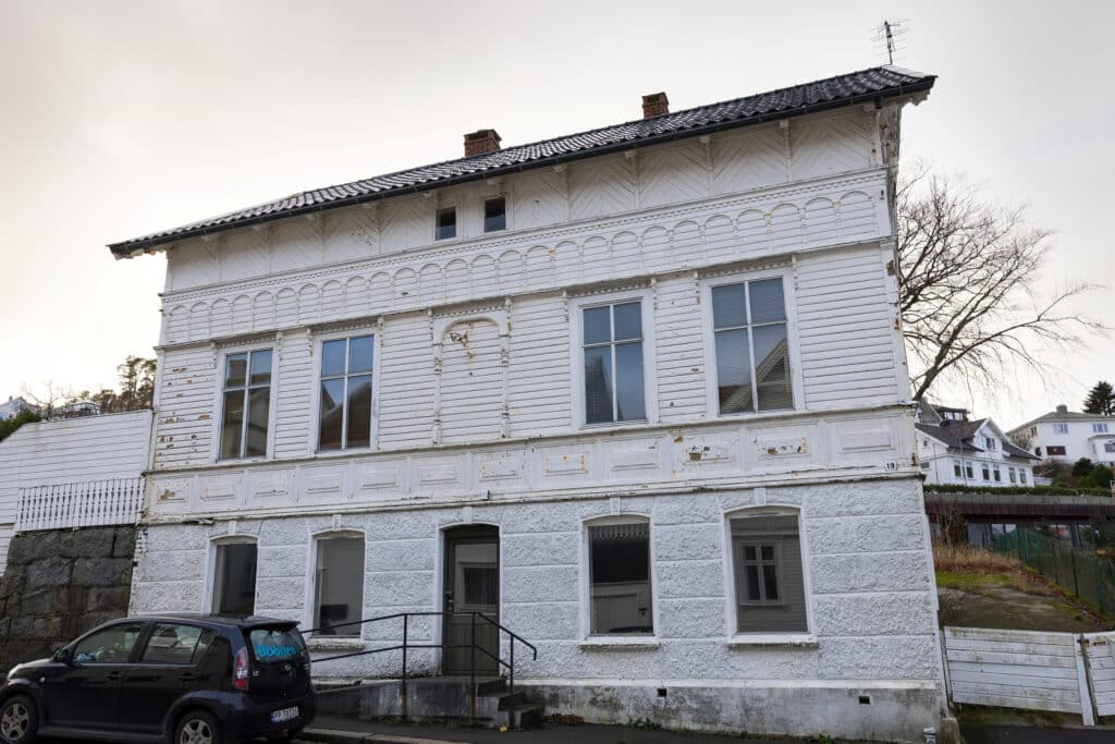 Et hvitt trehus i to etasjer med forvitret maling, flere store vinduer og en svart bil parkert foran på en overskyet dag.