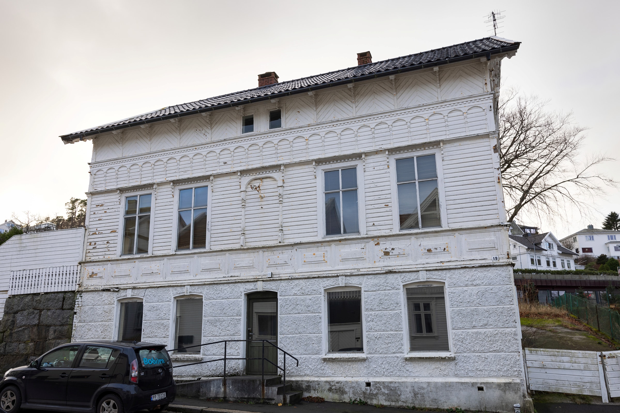 Et hvitt trehus i to etasjer med forvitret maling, flere store vinduer og en svart bil parkert foran på en overskyet dag.