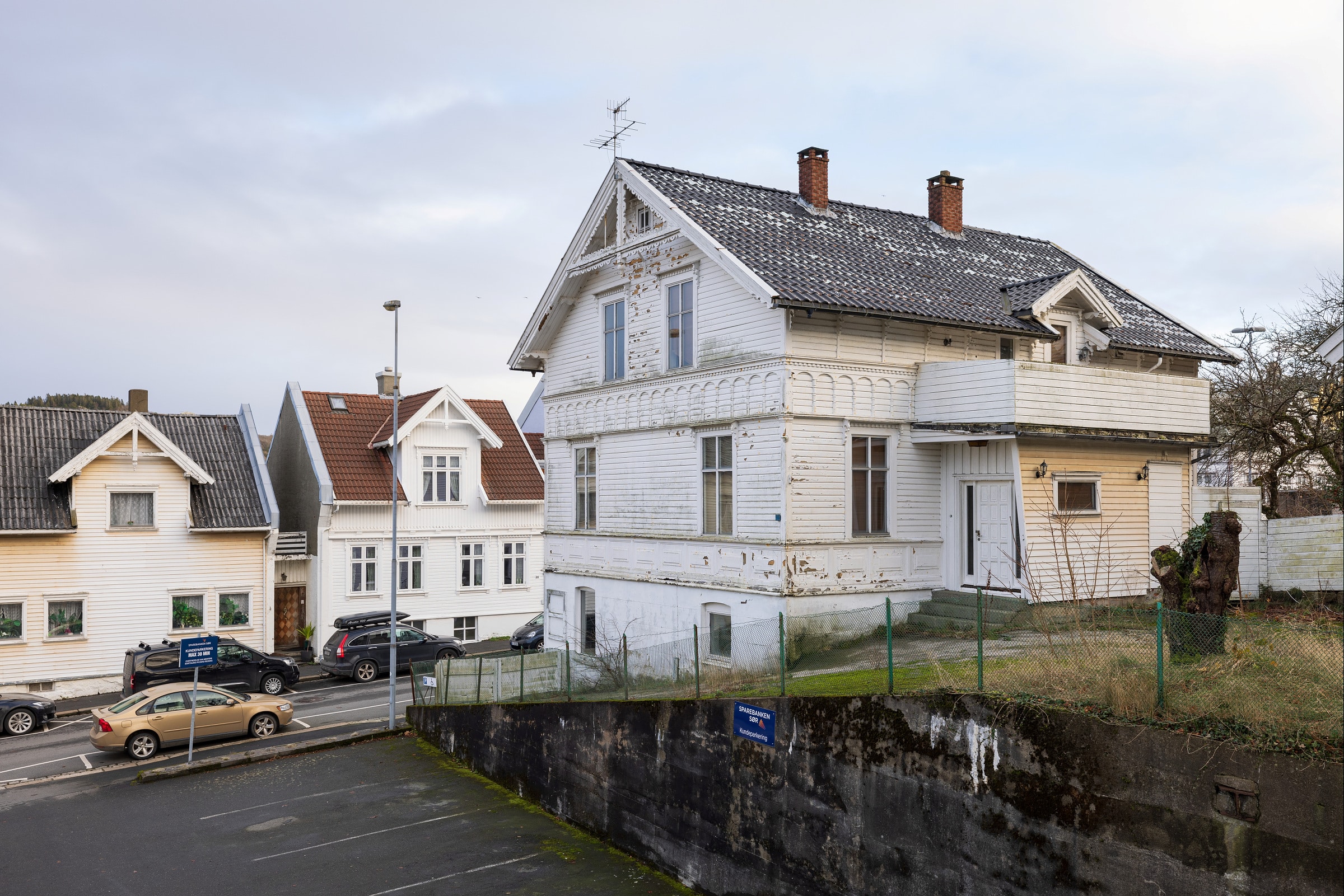 Et hvitt trehus i to etasjer med mørkt tak og noe avskallet maling, som ligger i en svak skråning ved siden av en gate og en parkeringsplass, med synlige nabohus.