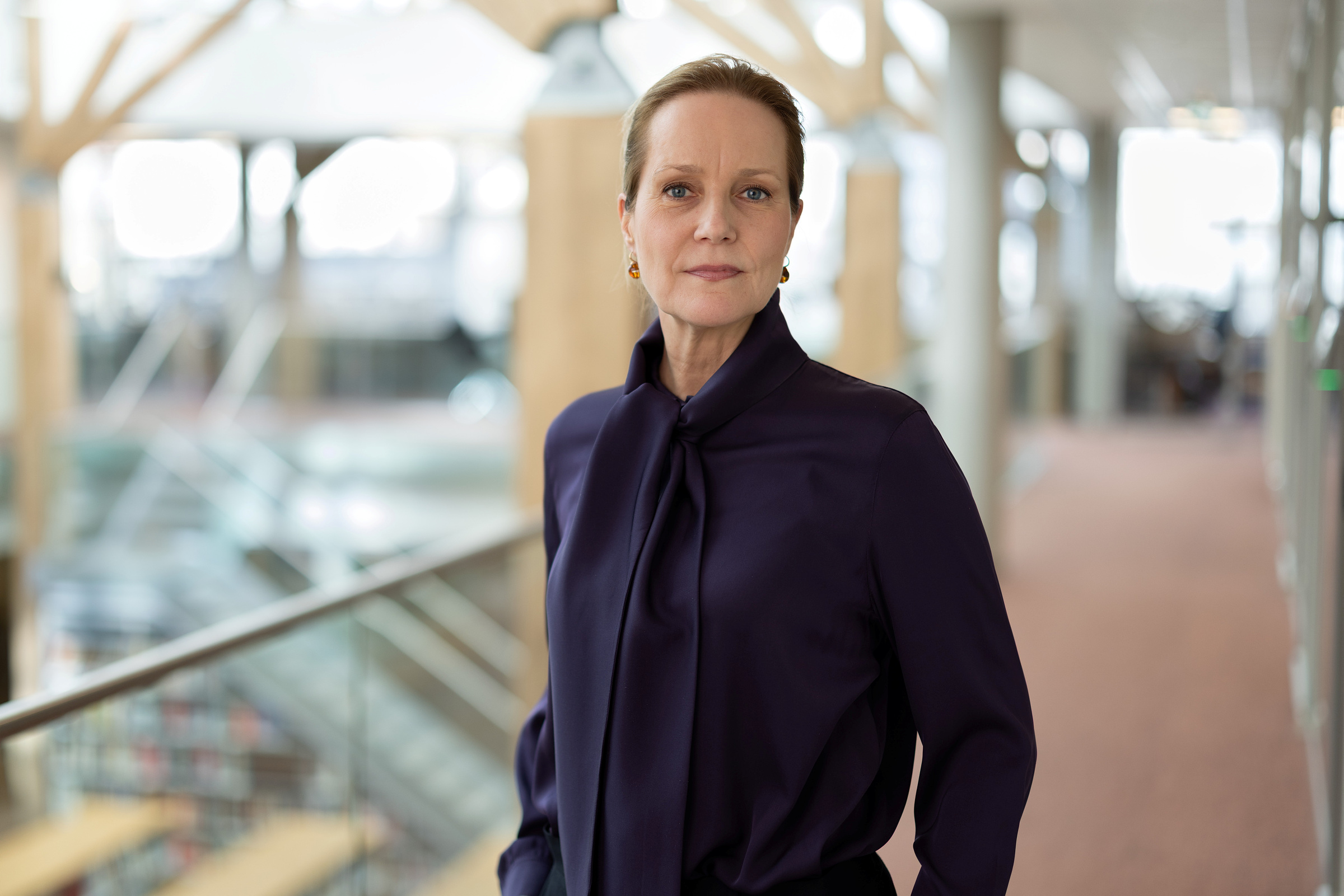 En kvinne i mørk bluse står i en moderne kontorkorridor og ser inn i kameraet med et nøytralt uttrykk. Bakgrunnen er ute av fokus.