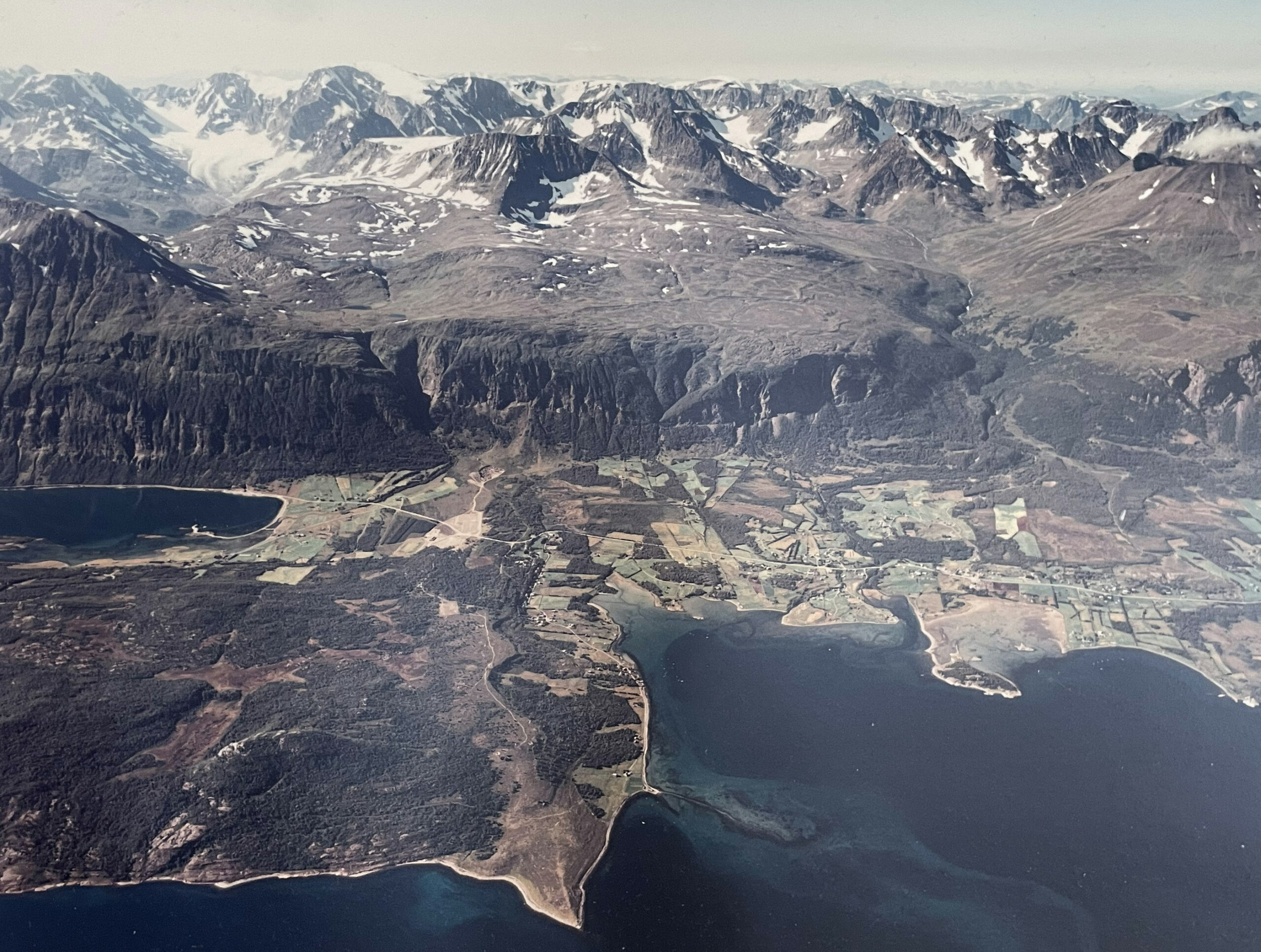 Gården Nedrum med Nergaardhuset 7 Flyfoto av et fjellandskap med snøflekker, skoger, åkrer og vannmasser, inkludert en kystlinje og innsjøer.