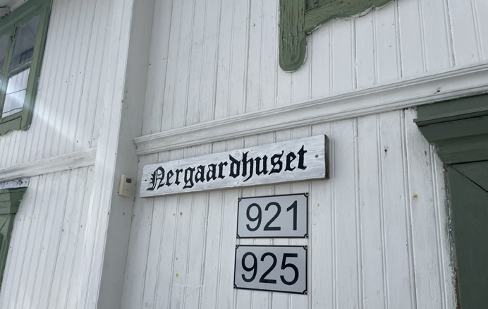 Gården Nedrum med Nergaardhuset 5 En hvit trebygning med grønne detaljer har et skilt med teksten Nergaardhuset og adressenumrene 921 og 925.