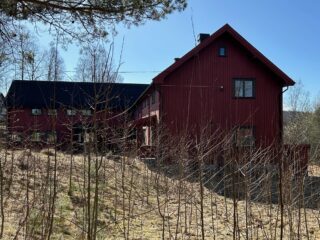 Et stort, rødt trehus med svart tak står i et gresskledd, skogkledd område under en klarblå himmel.