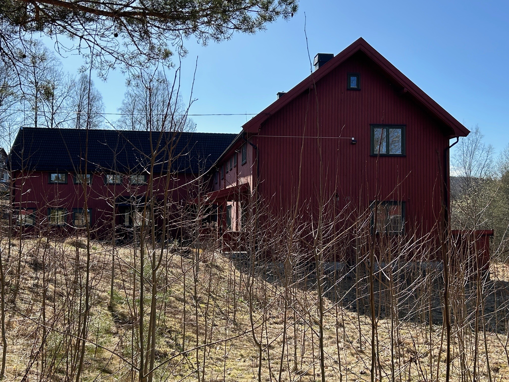 Et stort, rødt trehus med svart tak står i et gresskledd, skogkledd område under en klarblå himmel.