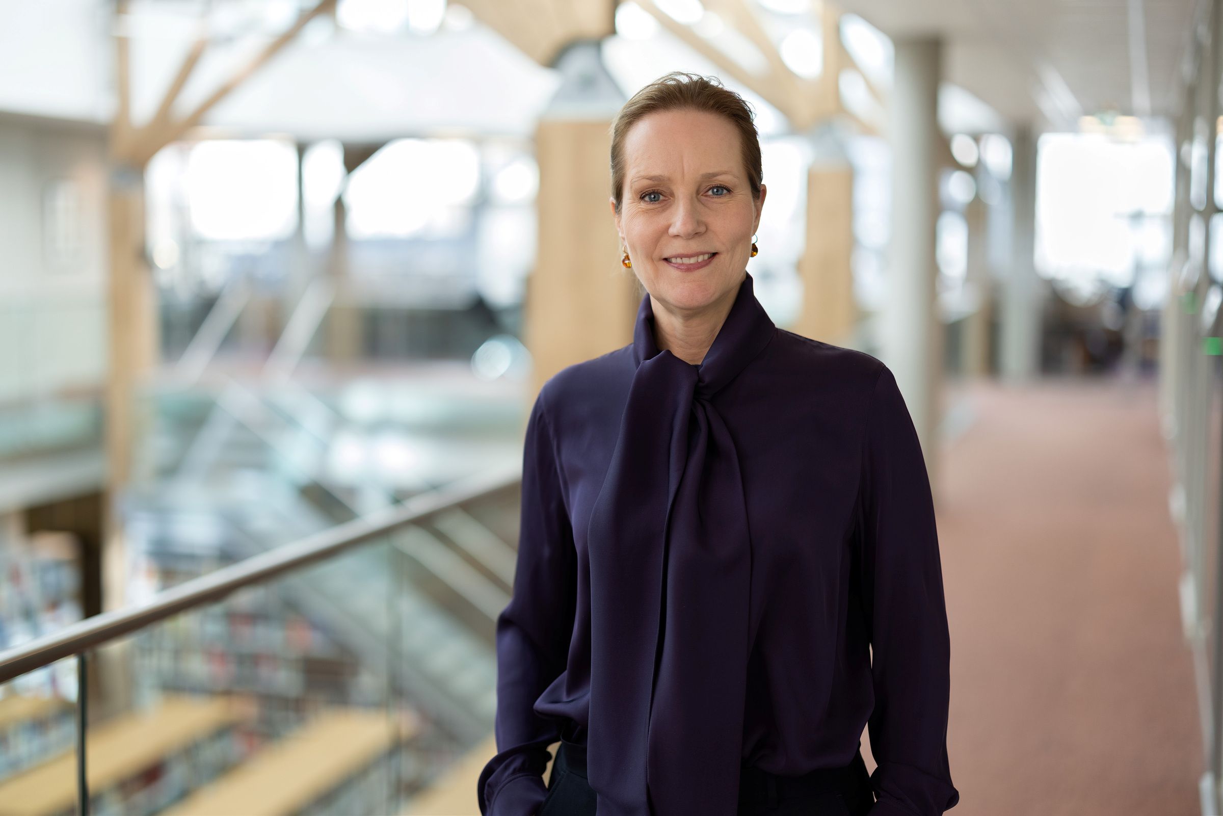 En kvinne i lilla bluse står innendørs i en moderne bygning med glassrekkverk og trebjelker og smiler til kameraet.