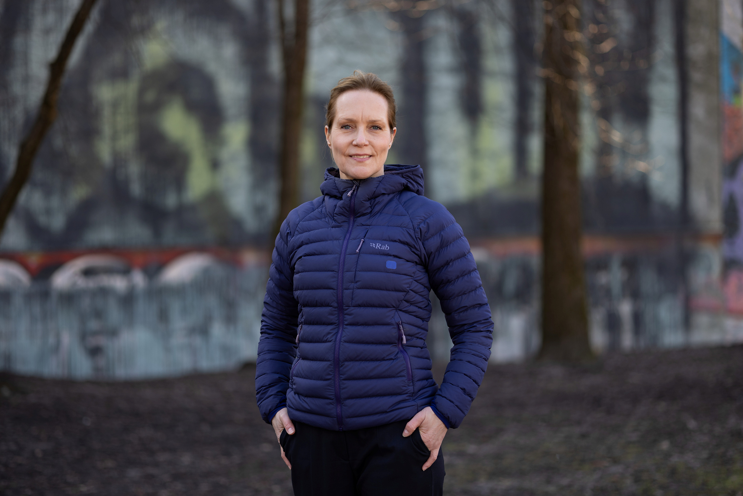 En kvinne i blå pufferjakke står utendørs med hendene i lommene, foran en vegg med graffiti og trær i bakgrunnen.