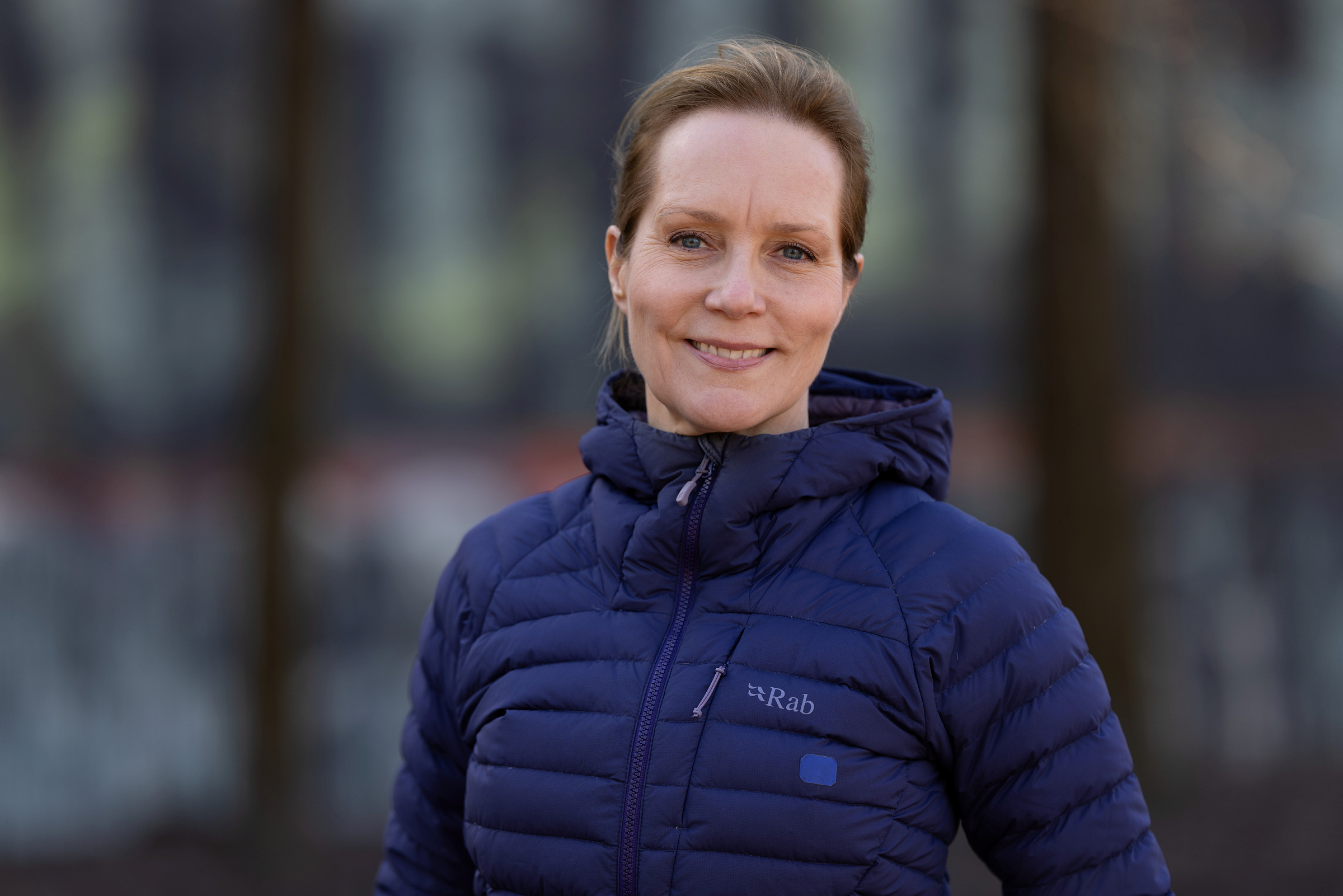 En kvinne i blå pufferjakke står utendørs og smiler til kameraet mot en uskarp bakgrunn.