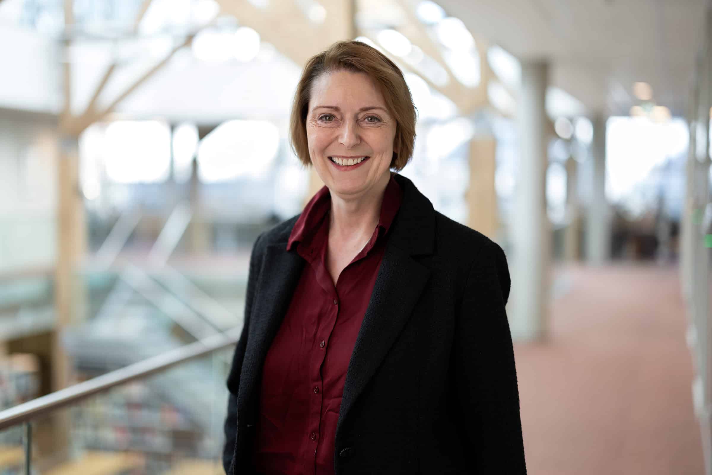 En kvinne med kort, lysebrunt hår, iført mørk blazer og rødbrun skjorte, står innendørs i en sterkt opplyst gang og smiler til kameraet.
