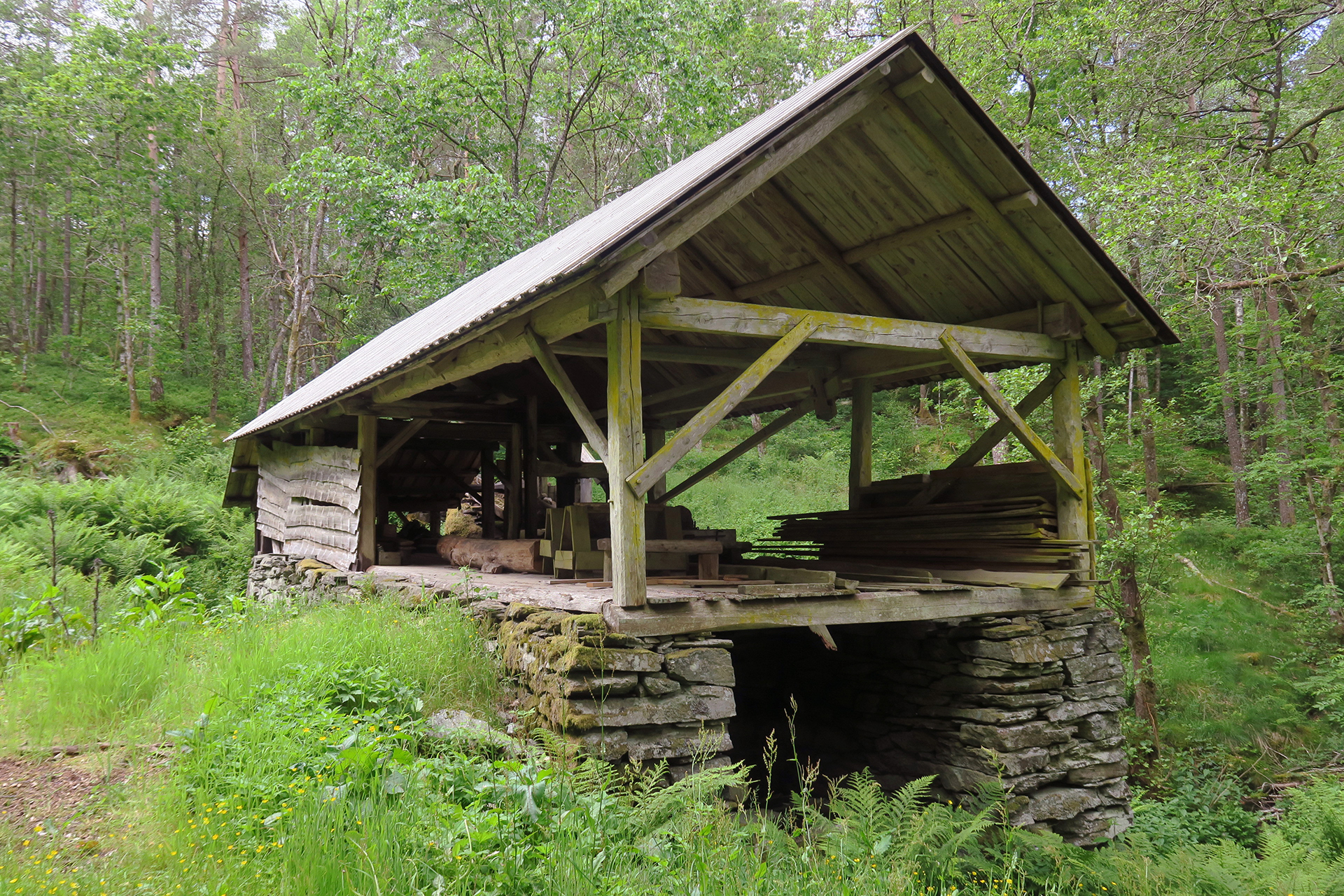 Et sagbruk i tre med skråtak står på et steinfundament i en skog, med stabler av tømmer og tømmerstokker synlig på innsiden.