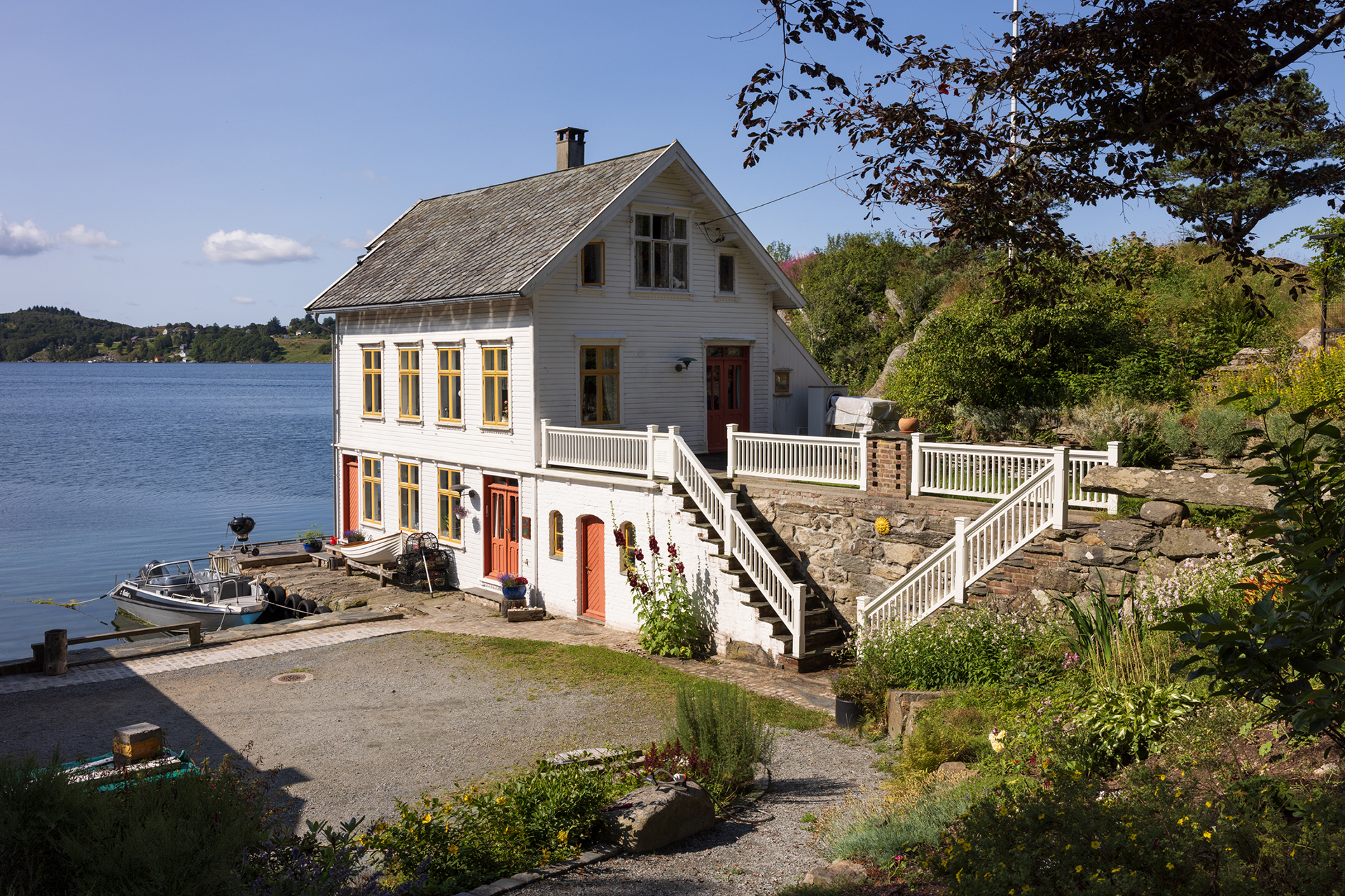 Et hvitt toetasjes hus med skråtak ligger ved et rolig vann, med en liten brygge og båt i nærheten, omgitt av grøntområder og en steinterrasse.