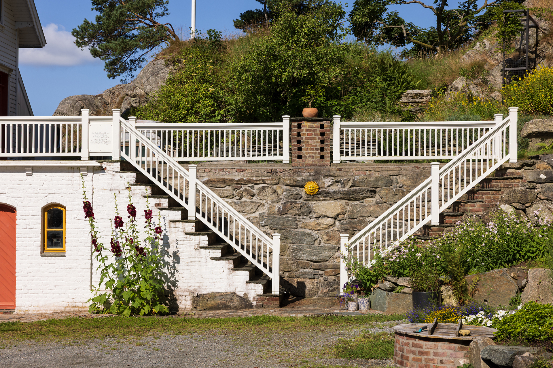 Utendørstrapp i stein og murstein med hvitt rekkverk, som fører opp til et terrassert hageområde; gule blomster og grønne planter omkranser trappen.