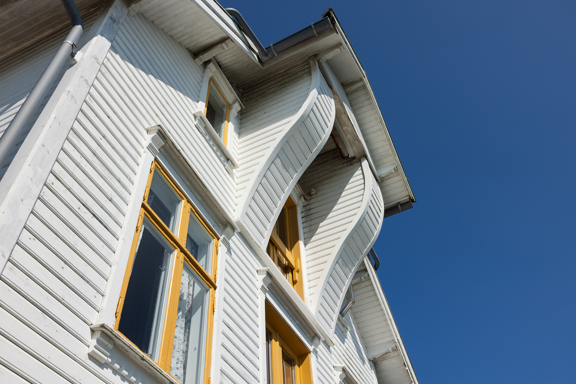 Et hvitt trehus med gule vinduer har skjeve, bølgende yttervegger under en klarblå himmel.