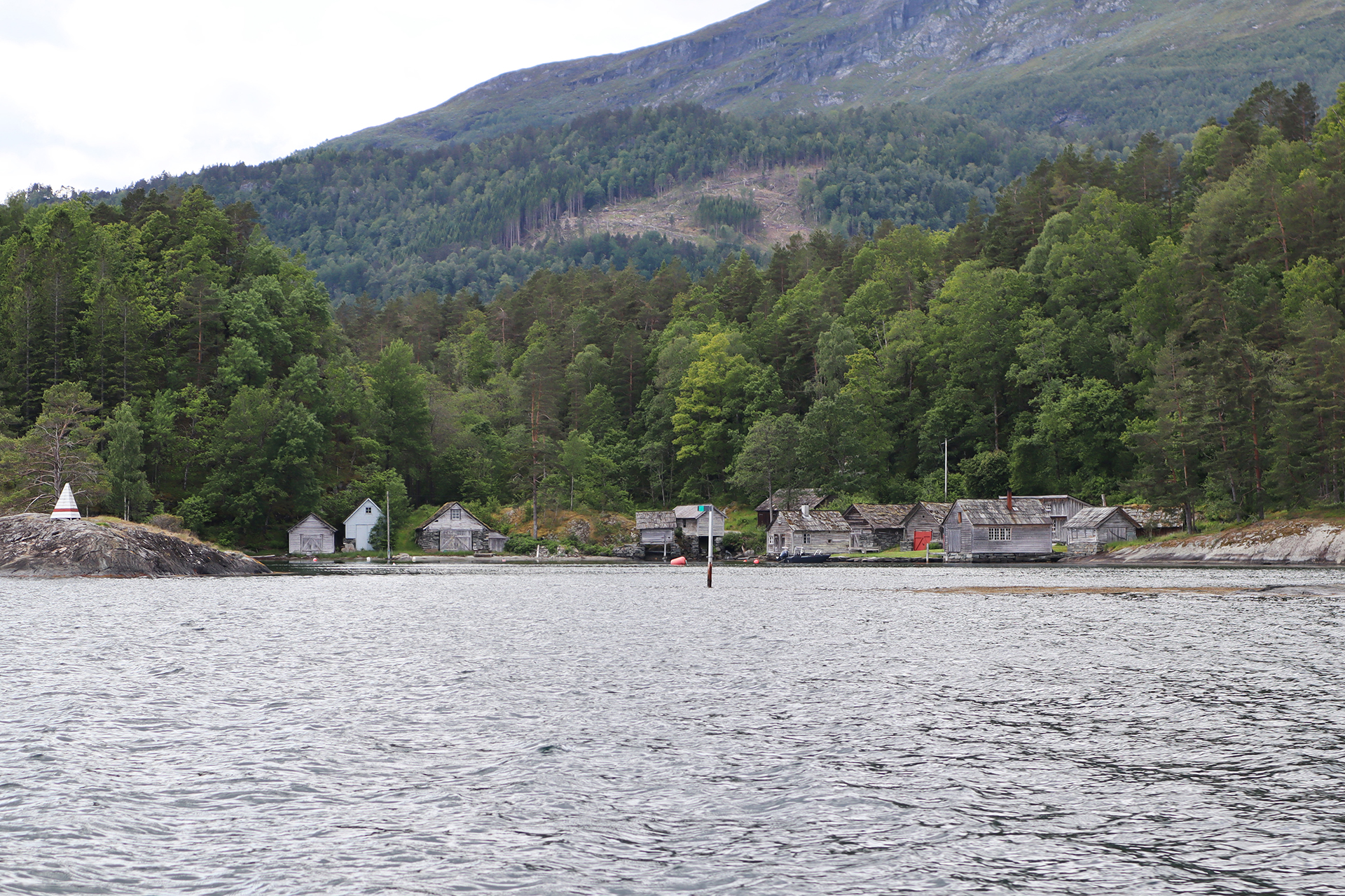 Små trehytter og hus ligger langs en innsjø eller fjord, med skogkledde åser i bakgrunnen.