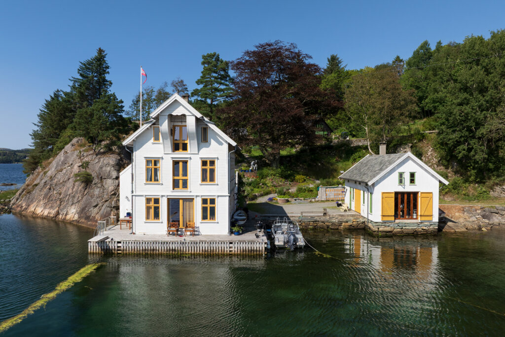 To hus ved vannkanten, ett stort og hvitt og ett mindre og gult, står ved siden av klart vann med trær og svaberg i bakgrunnen på en solrik dag.