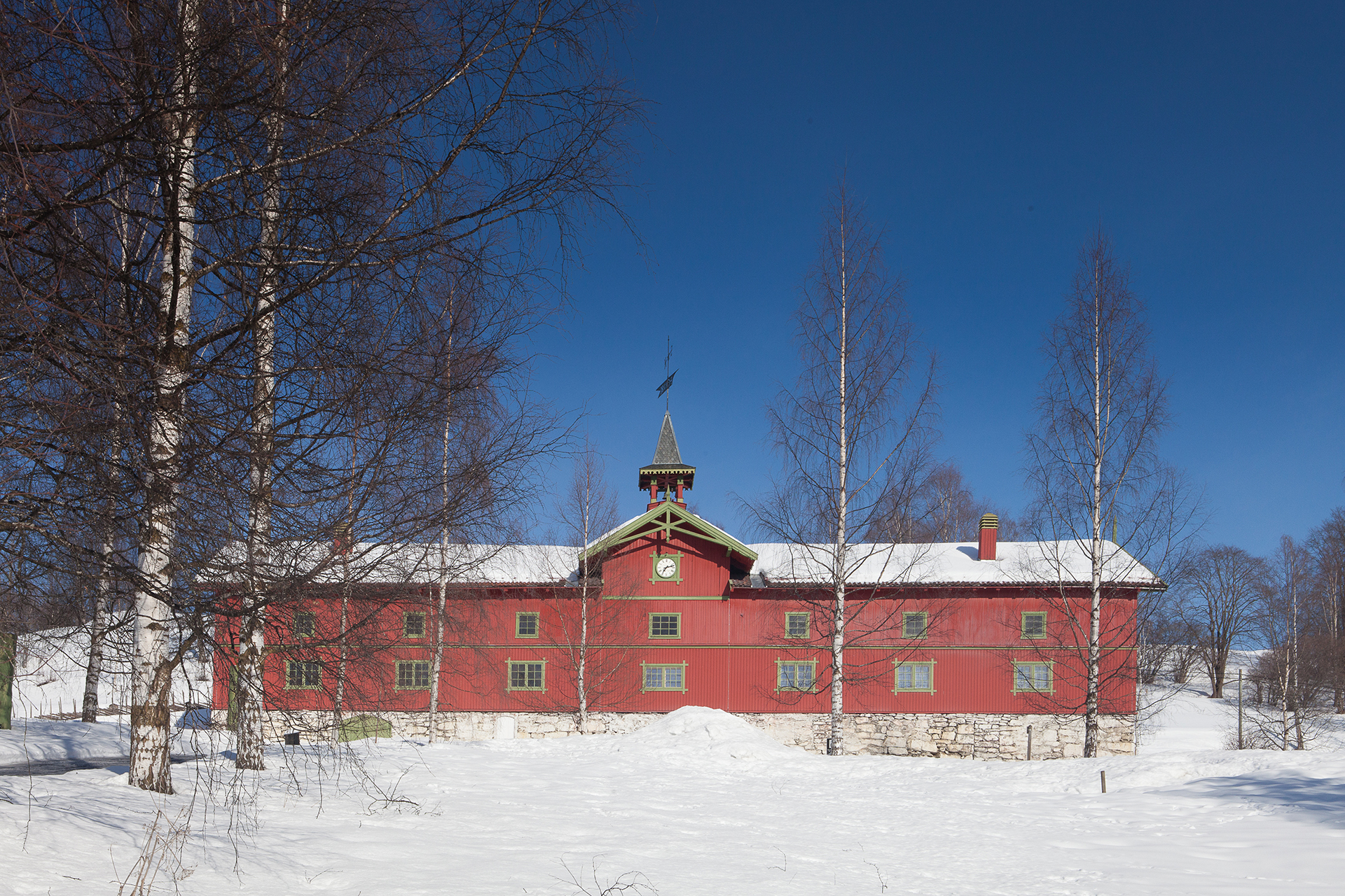 Får lov til å vere låve, men med ny bruk 6 En rød bygning med et sentralt klokketårn står i et snødekt landskap, omgitt av løvløse trær under en klarblå himmel.