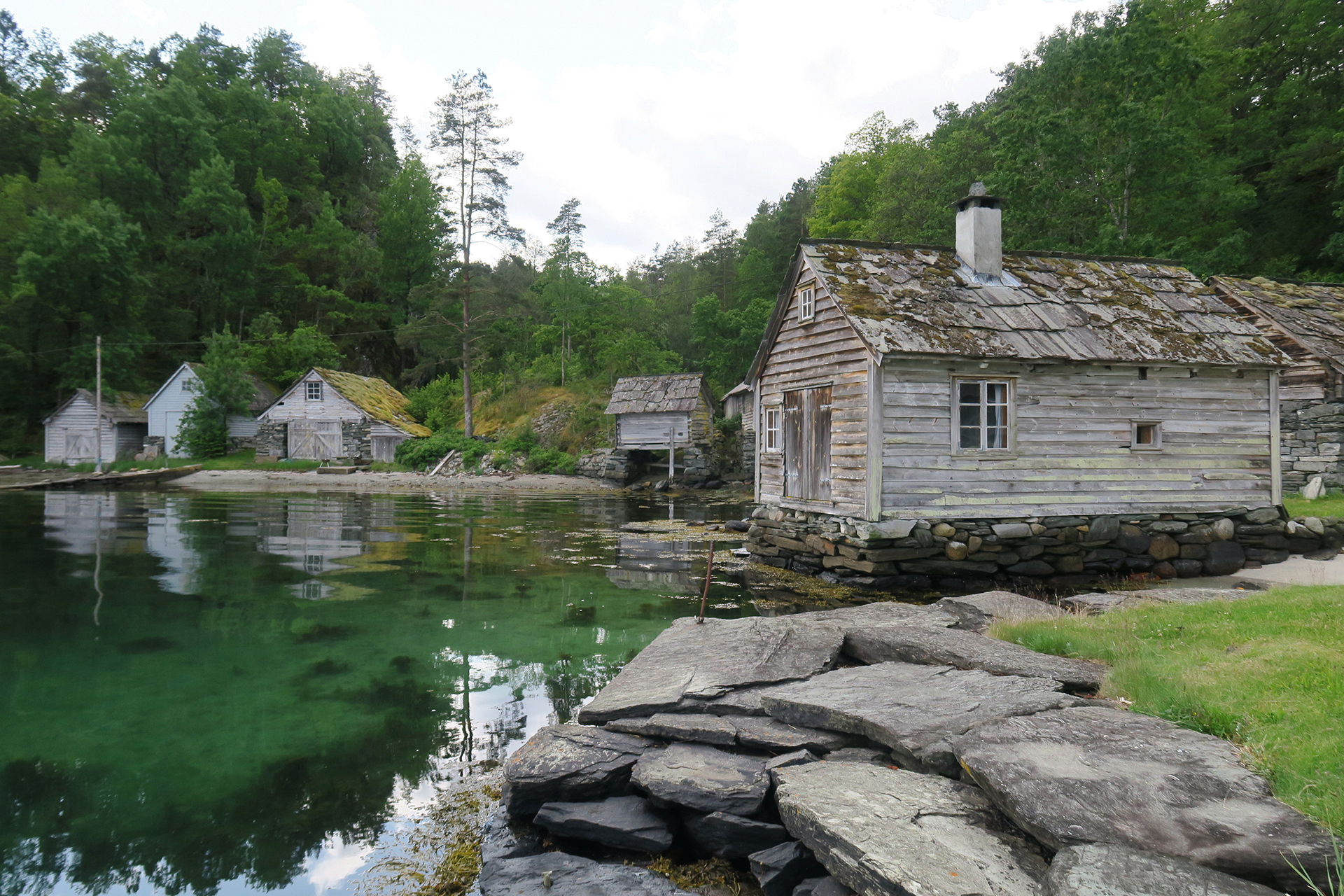 Gamle trehus med steinfundamenter står ved en rolig, klar innsjø omgitt av tett, grønn skog og en steinete strandlinje.