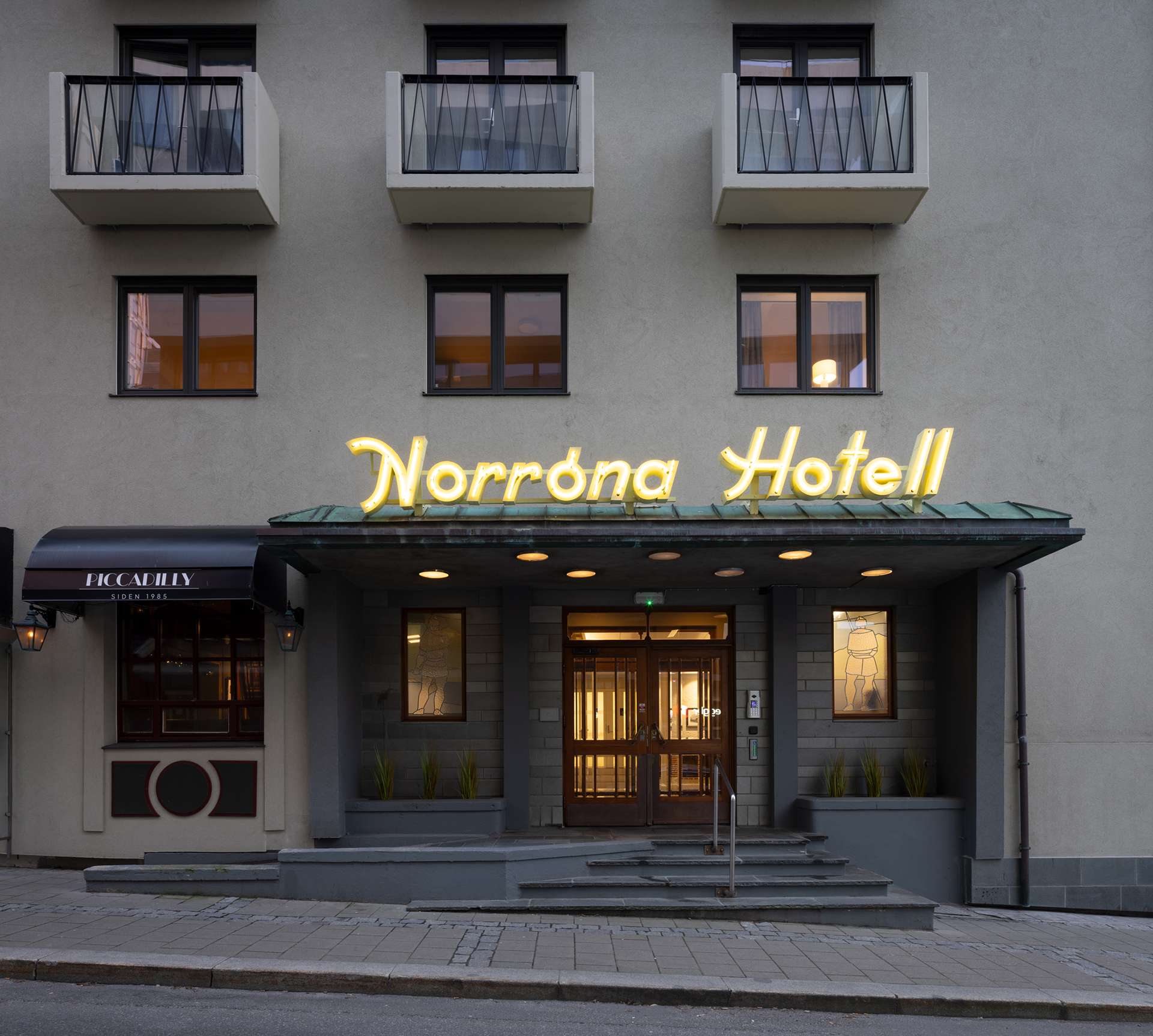 Fasaden på Norrøna Hotell med gul lysende skilting, tre balkonger over inngangen og en liten svart markise på venstre side.