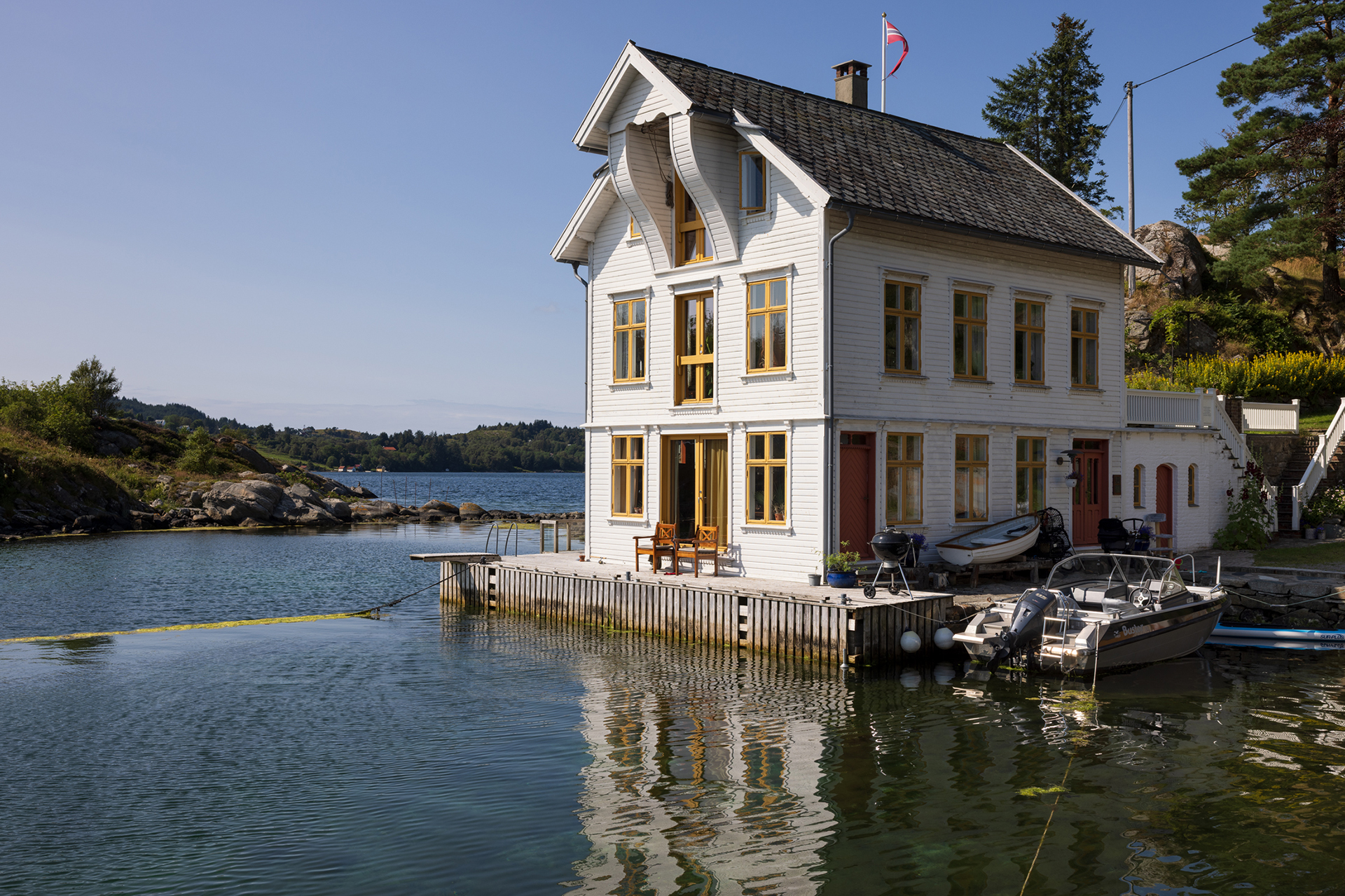 Et hvitt trehus med gule kanter ligger i utkanten av en rolig vannkant; båter ligger ved kai i nærheten, og i bakgrunnen skimtes trær under en klar himmel.