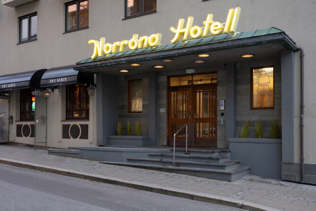 Inngangspartiet til Norrøna Hotell med neonskilt, grønn markise, glassdører og to opplyste glassmalerier på hver side, beliggende i en stille gate.