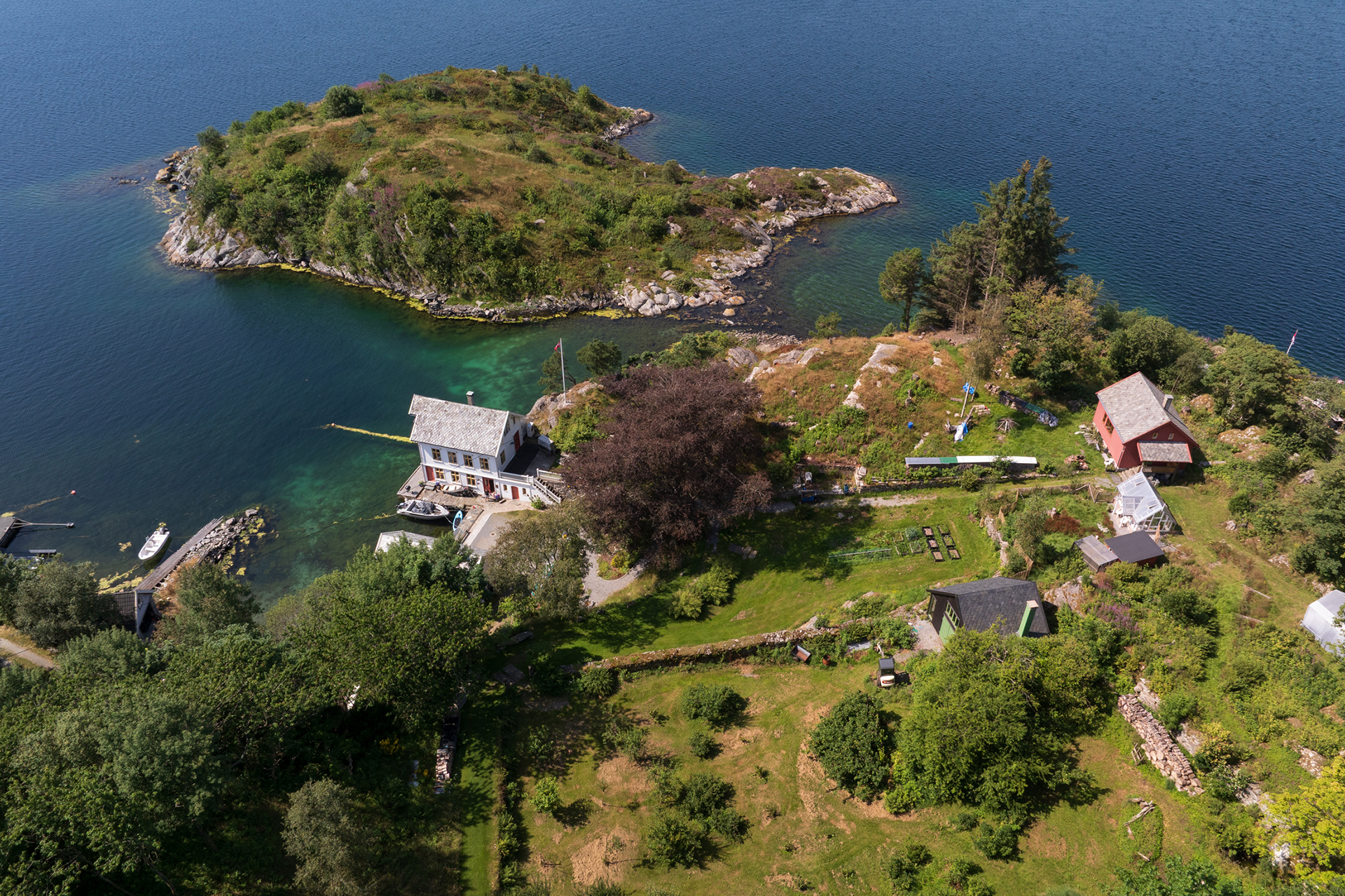 Flyfoto av en landlig eiendom ved en innsjø med flere hus, frodige grøntområder, hager og en liten øy i vannet.
