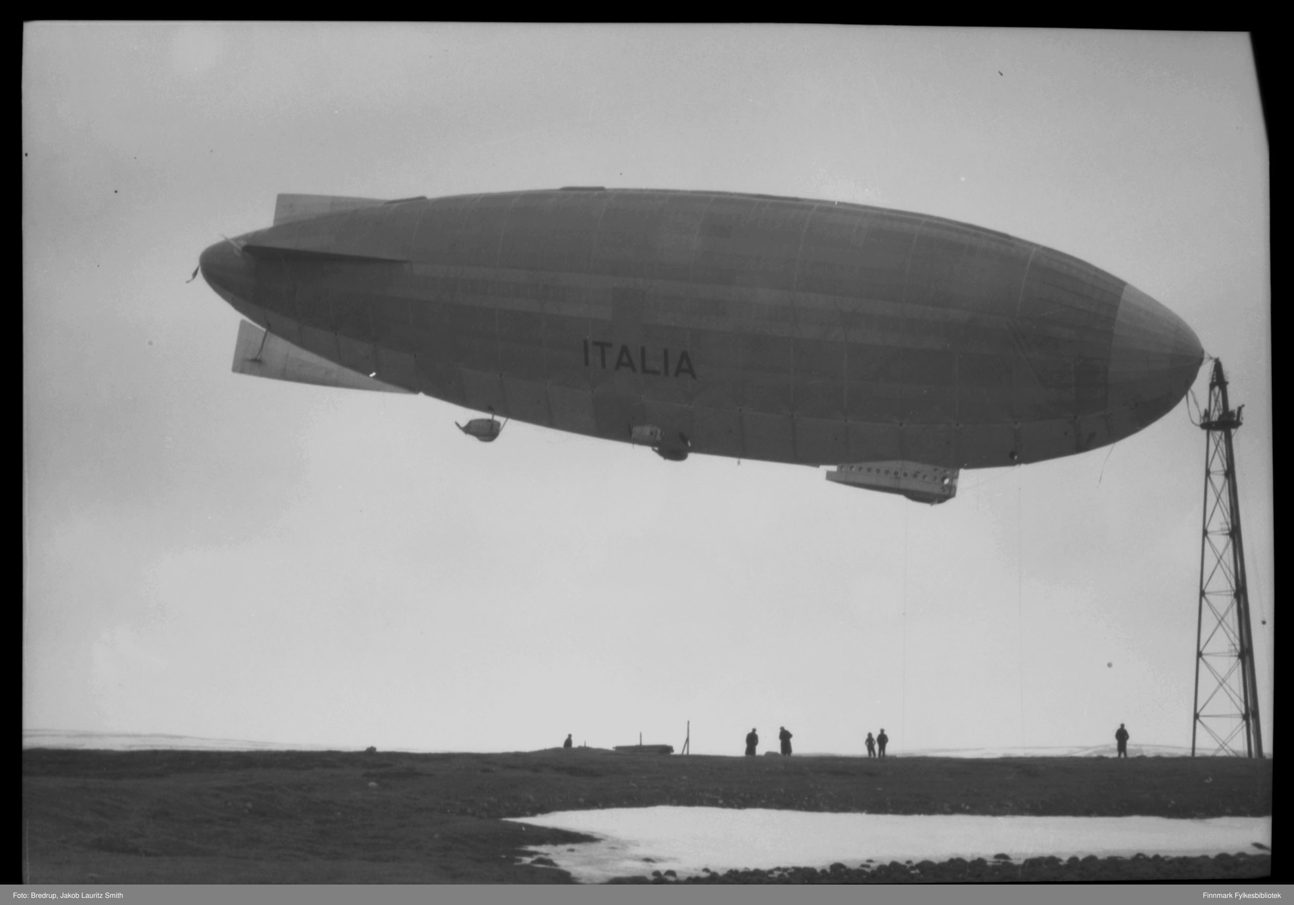 Svart-hvitt-foto av luftskipet Italia som flyr lavt over et snødekt, flatt landskap med flere mennesker og en høy bygning under seg.