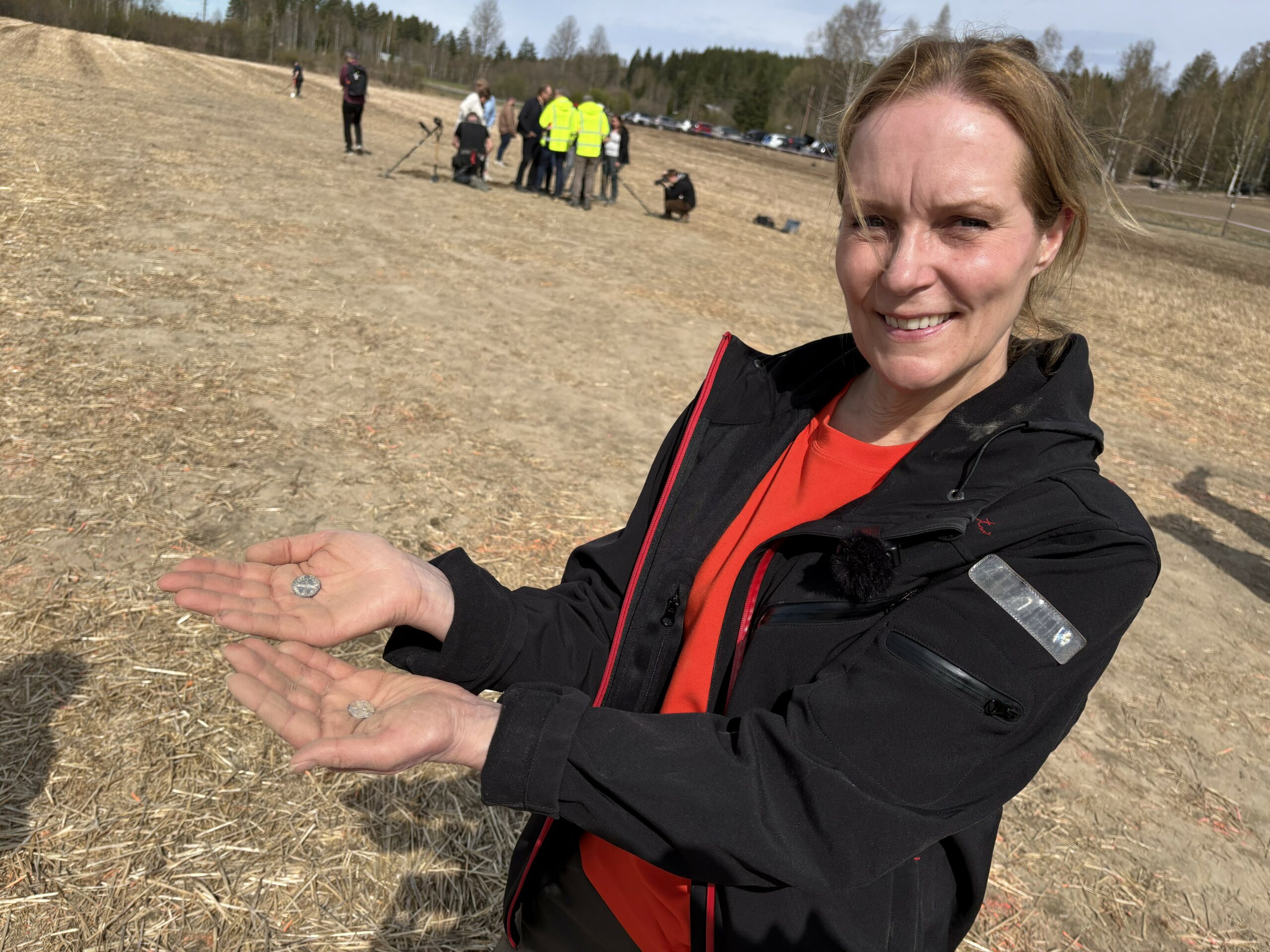 En kvinne står på et jorde og holder små gjenstander i hendene, mens en gruppe mennesker er synlige i bakgrunnen.
