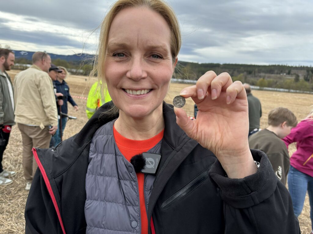 En kvinne står utendørs på et jorde og holder opp en mynt, mens hun smiler til kameraet, med flere personer i bakgrunnen.