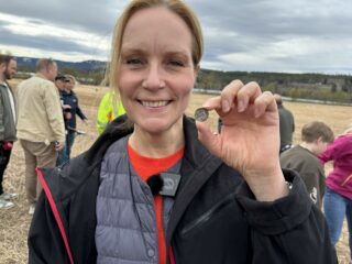 En kvinne står utendørs på et jorde og holder opp en mynt, mens hun smiler til kameraet, med flere personer i bakgrunnen.